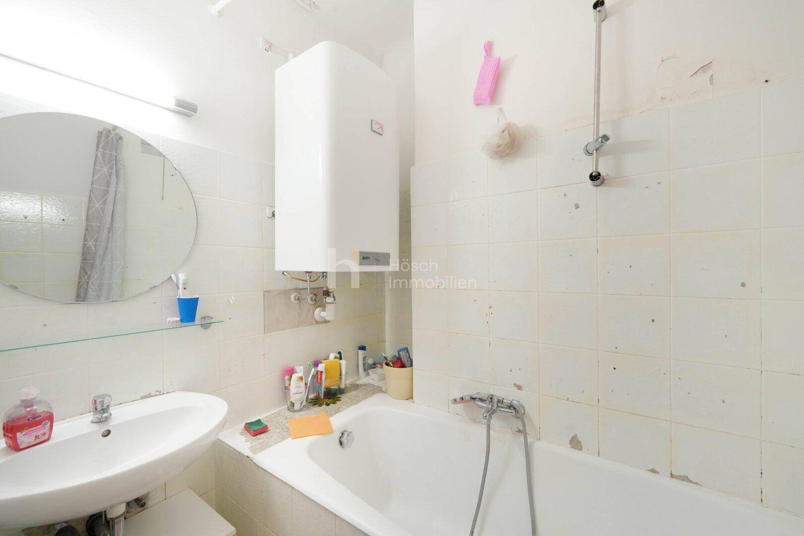 Badezimmer mit Badewanne, Waschbecken, Boiler und älteren Fliesen an den Wänden.