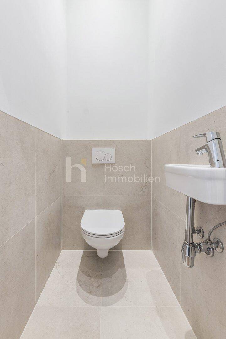 Separates WC mit wandhängender Toilette und kleinem Handwaschbecken, hell gefliest und funktional.