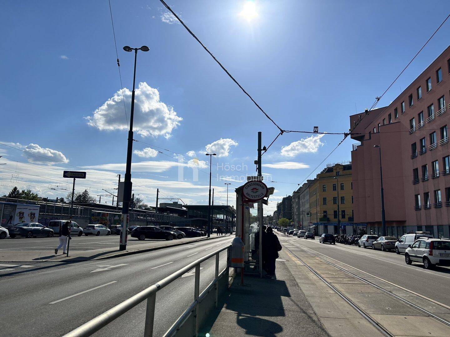 Sonnige Straßenansicht mit Straßenbahnschienen, Haltestelle und städtischer Bebauung im Hintergrund.