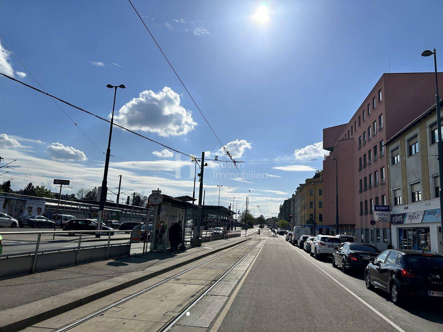 Weitläufige Straßenansicht an einem sonnigen Tag mit Straßenbahnschienen und städtischer Architektur.