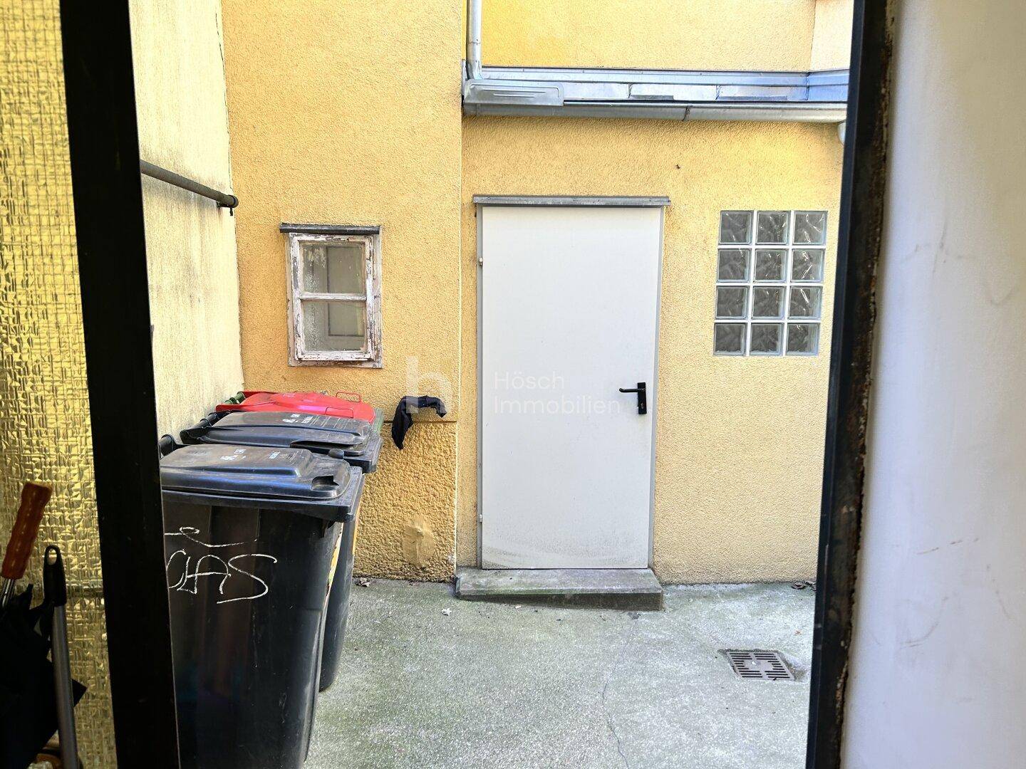 Schmaler Innenhof mit einer weißen Tür, kleinen Fenstern und Mülltonnen im Vordergrund.