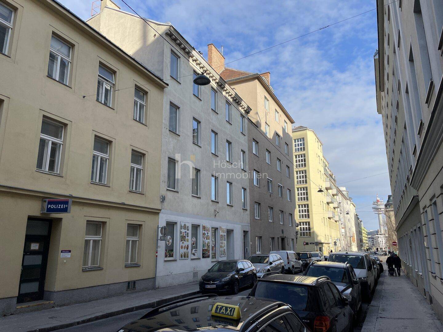 Straßenansicht mit parkenden Autos und Gebäuden unter einem blauen Himmel.