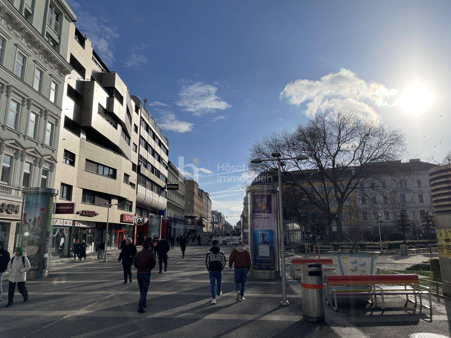 Belebtes Stadtbild mit Fußgängern, Geschäften und Gebäuden unter blauem Himmel.