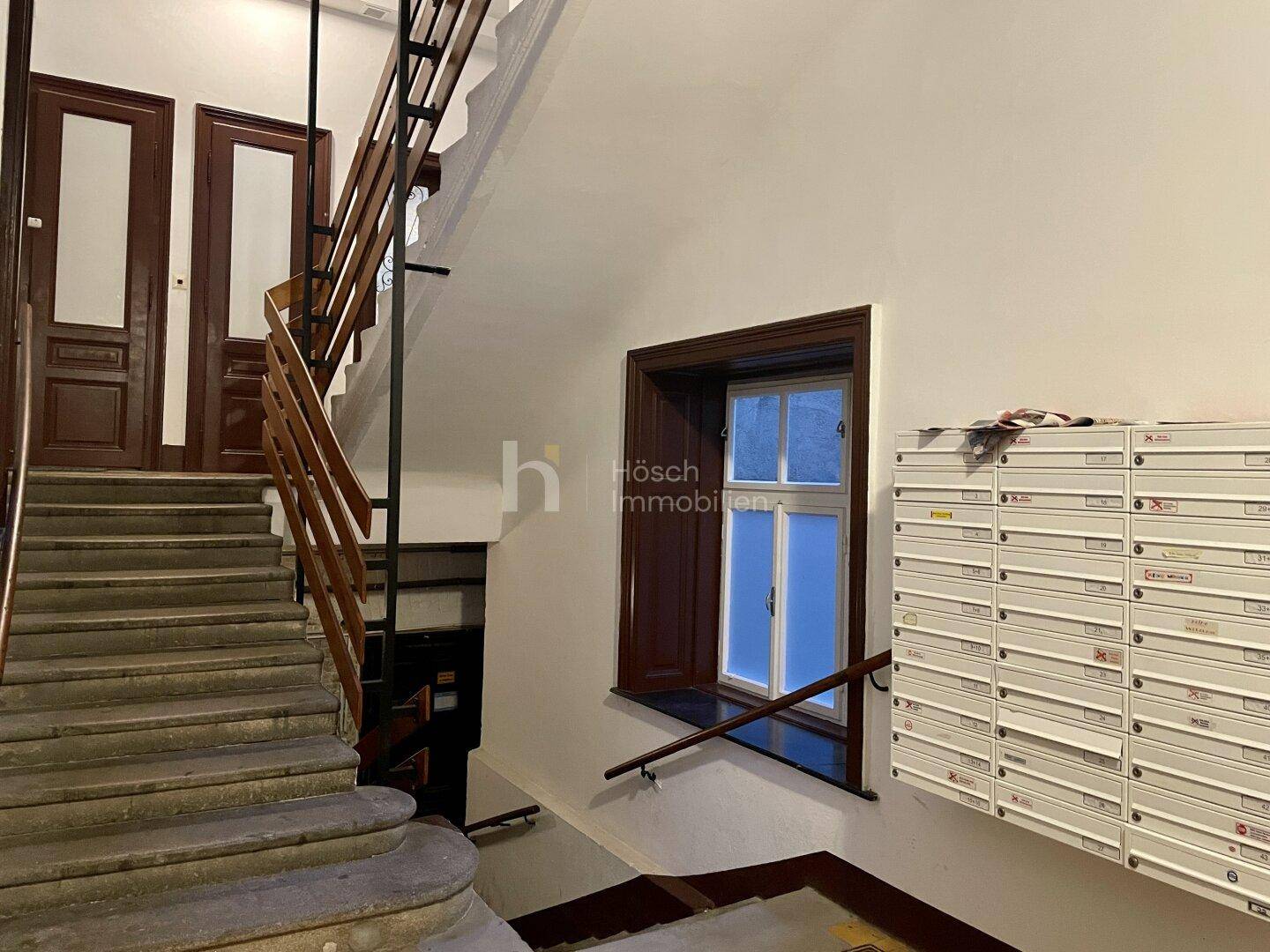 Treppenhaus eines Altbaus mit Holzgeländer, Steinstufen und Briefkästen an der Wand.