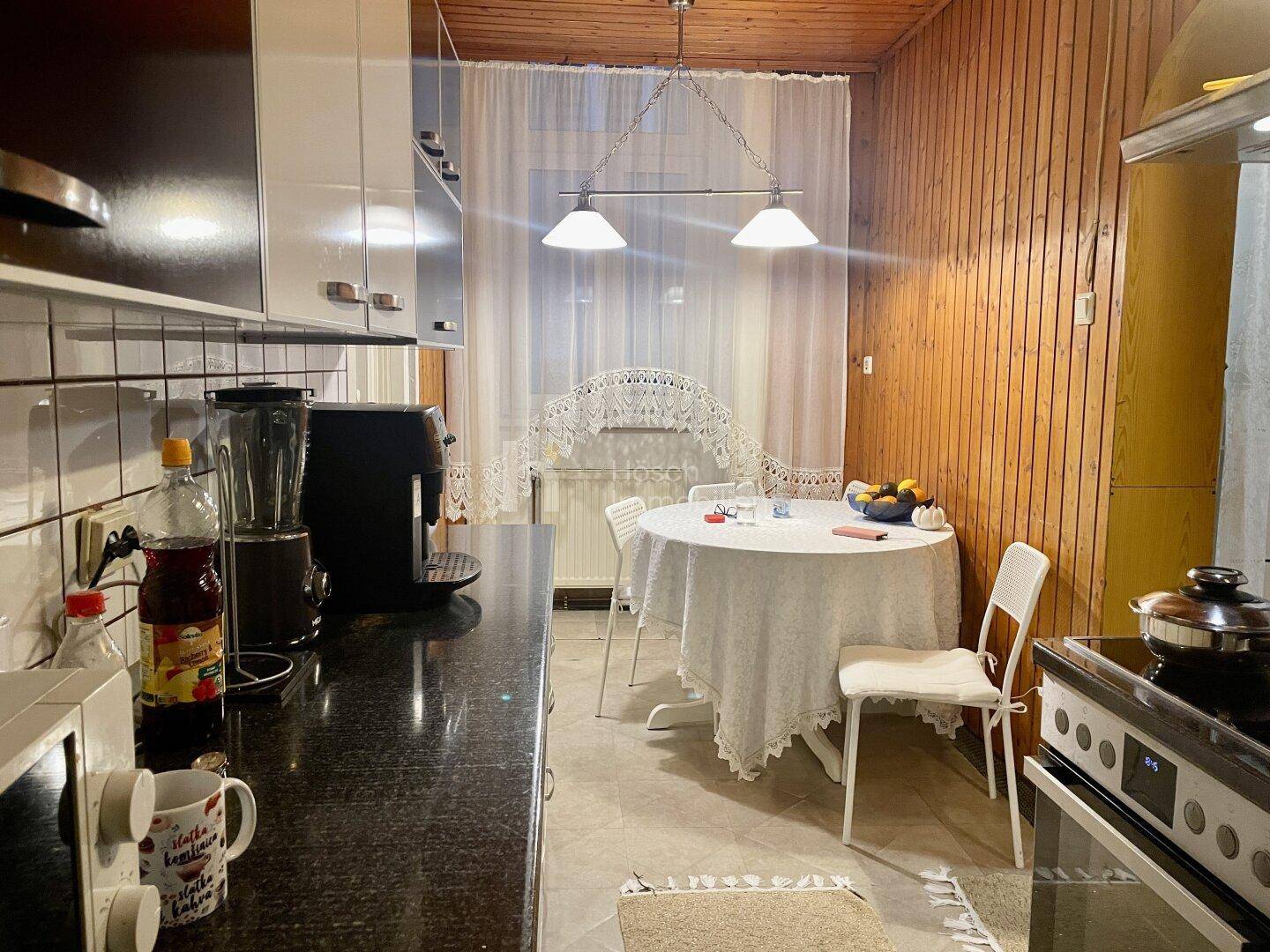 Küche mit dunkler Arbeitsplatte, Holzvertäfelung und einem kleinen Esstisch am Fenster.