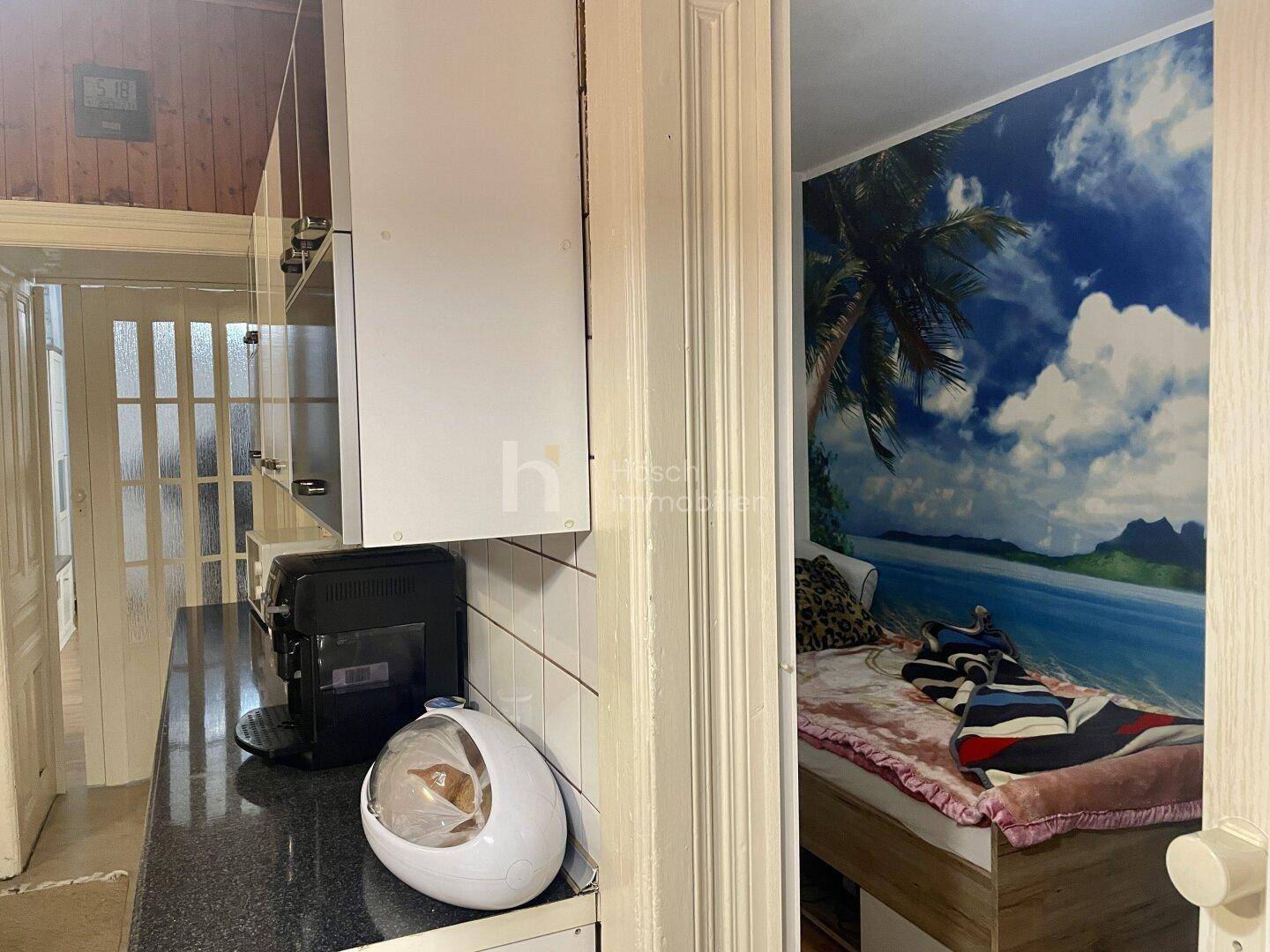 Küchenzeile mit Geräten und Blick in ein angrenzendes Zimmer mit tropischer Wandtapete.