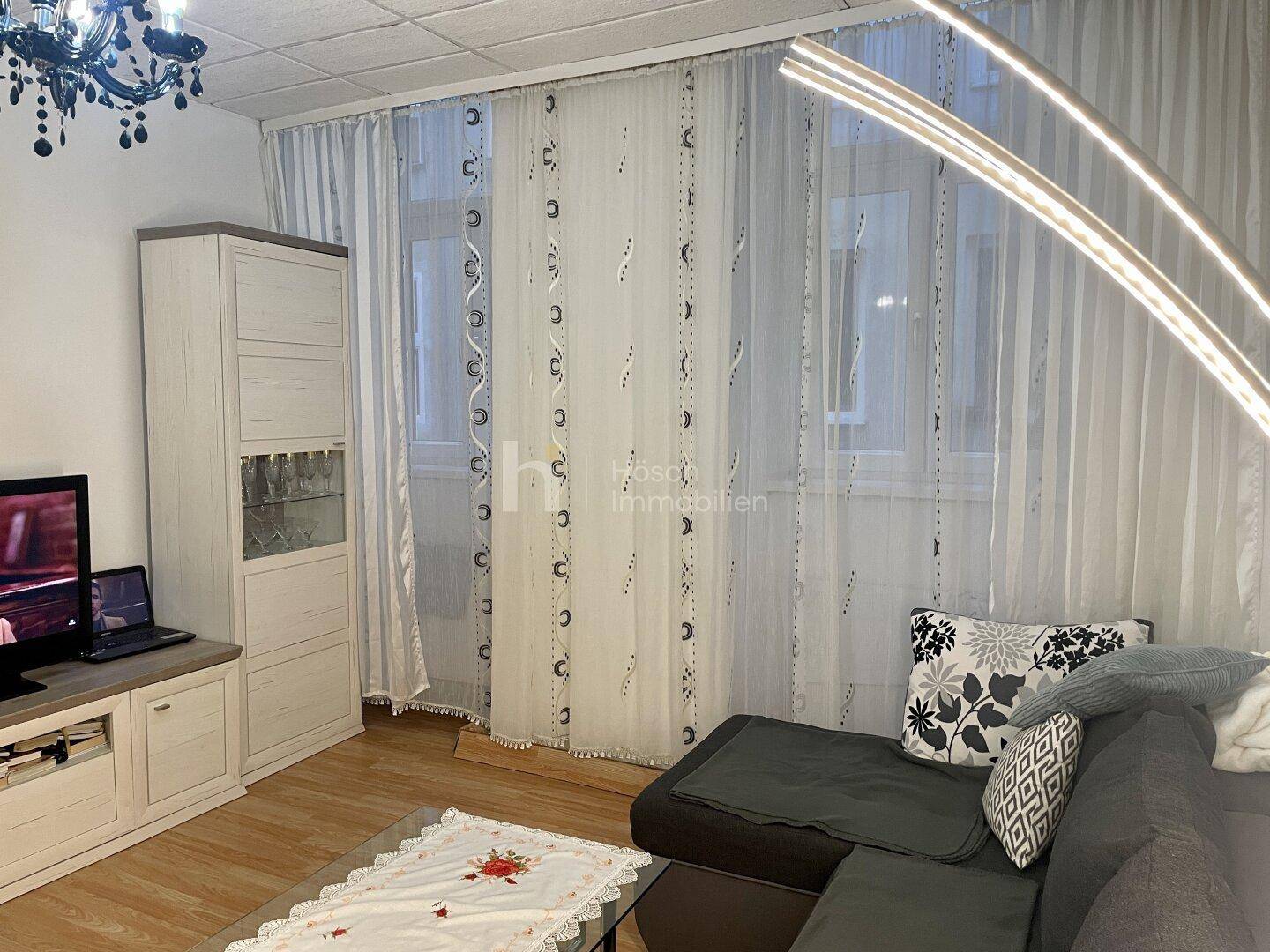 Gemütliches Wohnzimmer mit Sofa, Fernseher und hellen Vorhängen vor den Fenstern.