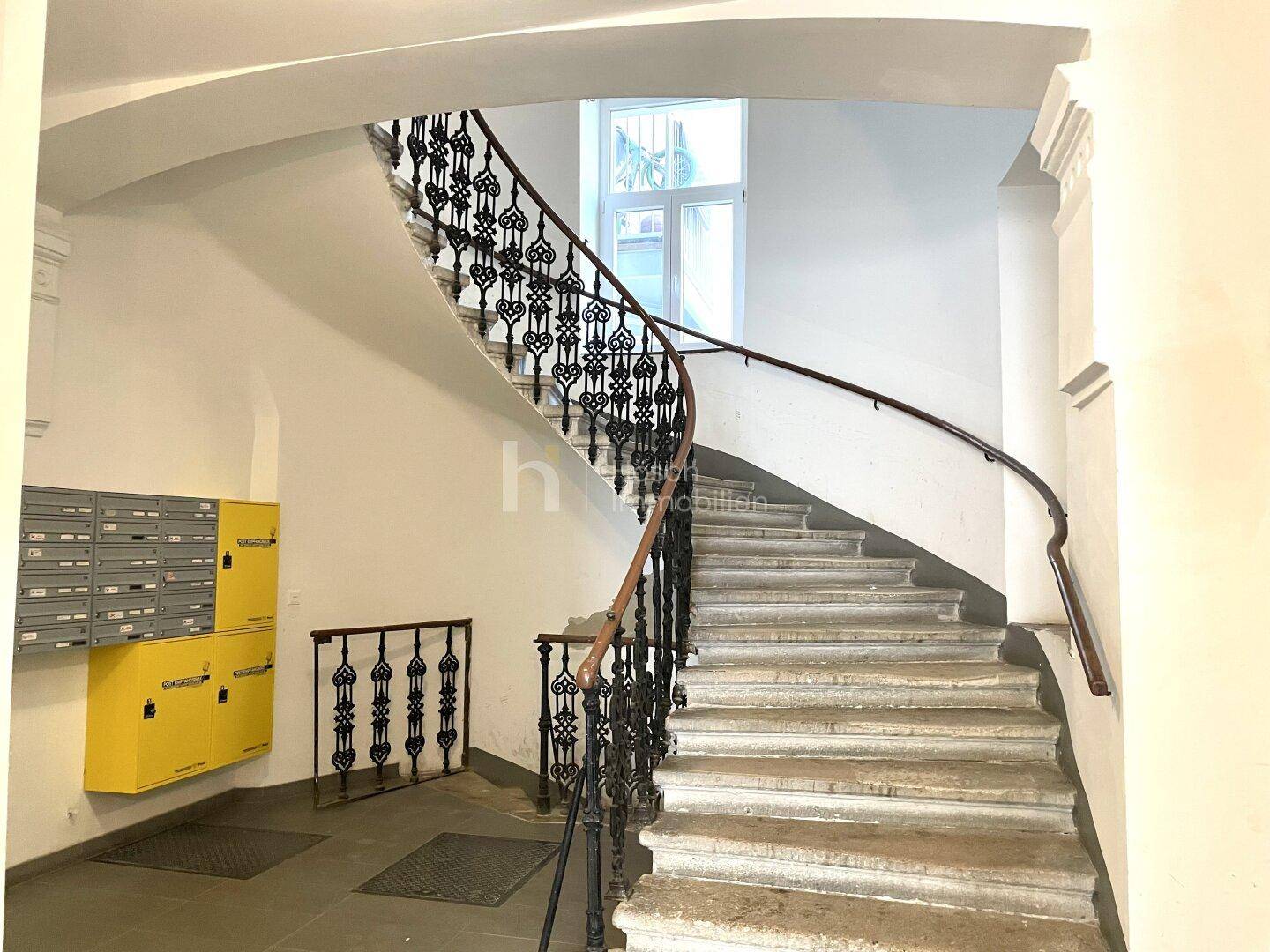 Elegantes Treppenhaus mit geschwungenem schmiedeeisernem Geländer und Steinstufen, beleuchtet durch ein Fenster.