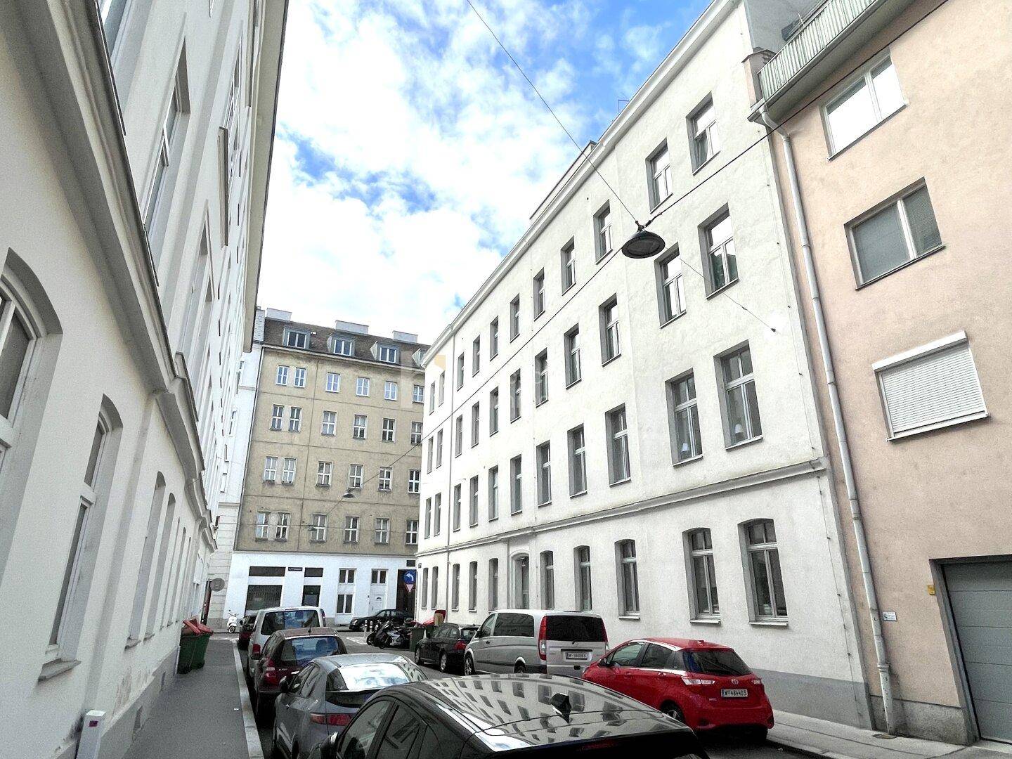 Enge Stadtstraße mit parkenden Autos und mehrstöckigen Gebäuden unter einem bewölkten Himmel.