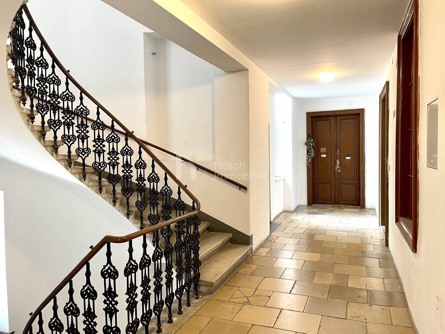 Geräumiger Flur mit Steinfliesenboden, einer eleganten Treppe und einer klassischen Holztür am Ende.