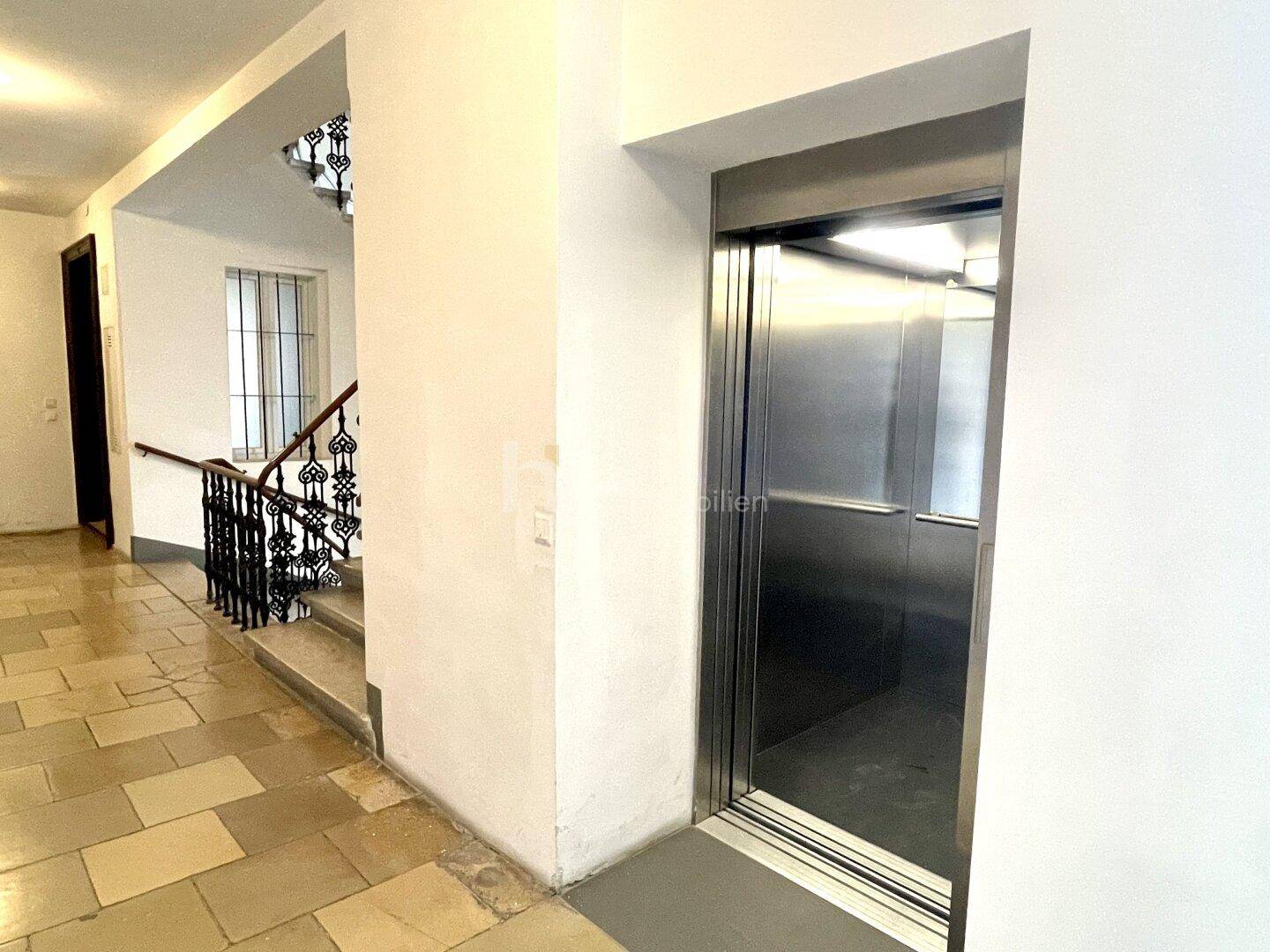 Moderner Aufzug mit Edelstahltüren neben einem Treppenaufgang in einem Mehrfamilienhaus.