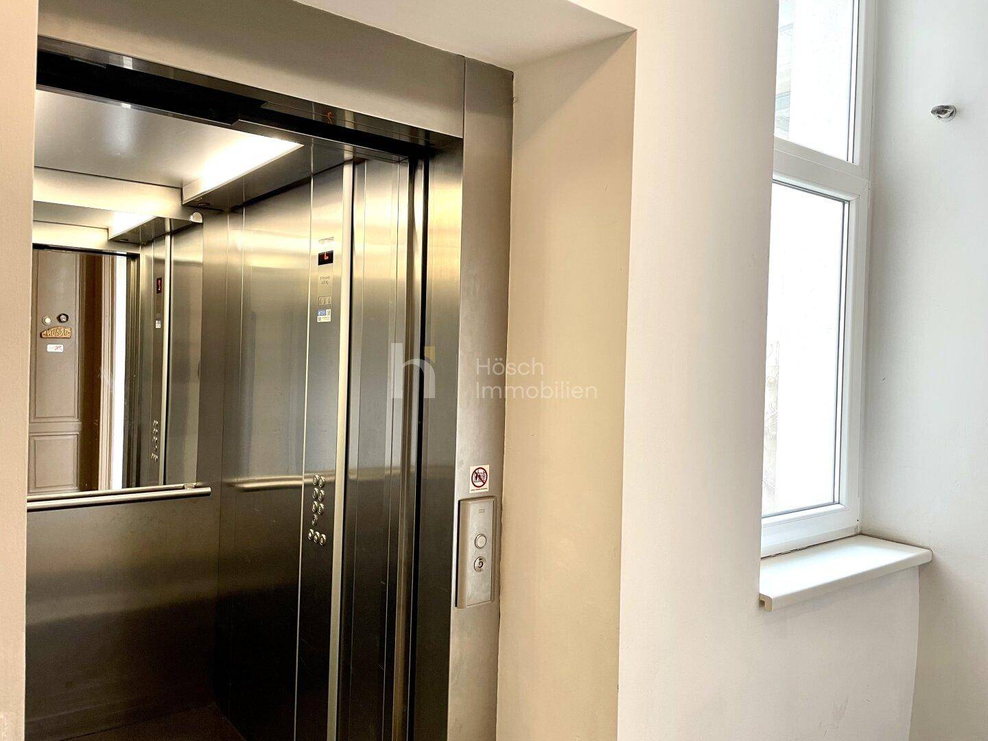 Detailansicht eines modernen Aufzugs mit Edelstahlverkleidung und Bedienfeld neben einem Fenster.
