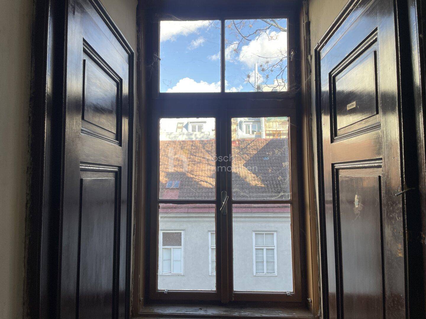 Blick aus einem Fenster mit Holzrahmen und Fensterläden auf gegenüberliegende Dächer.