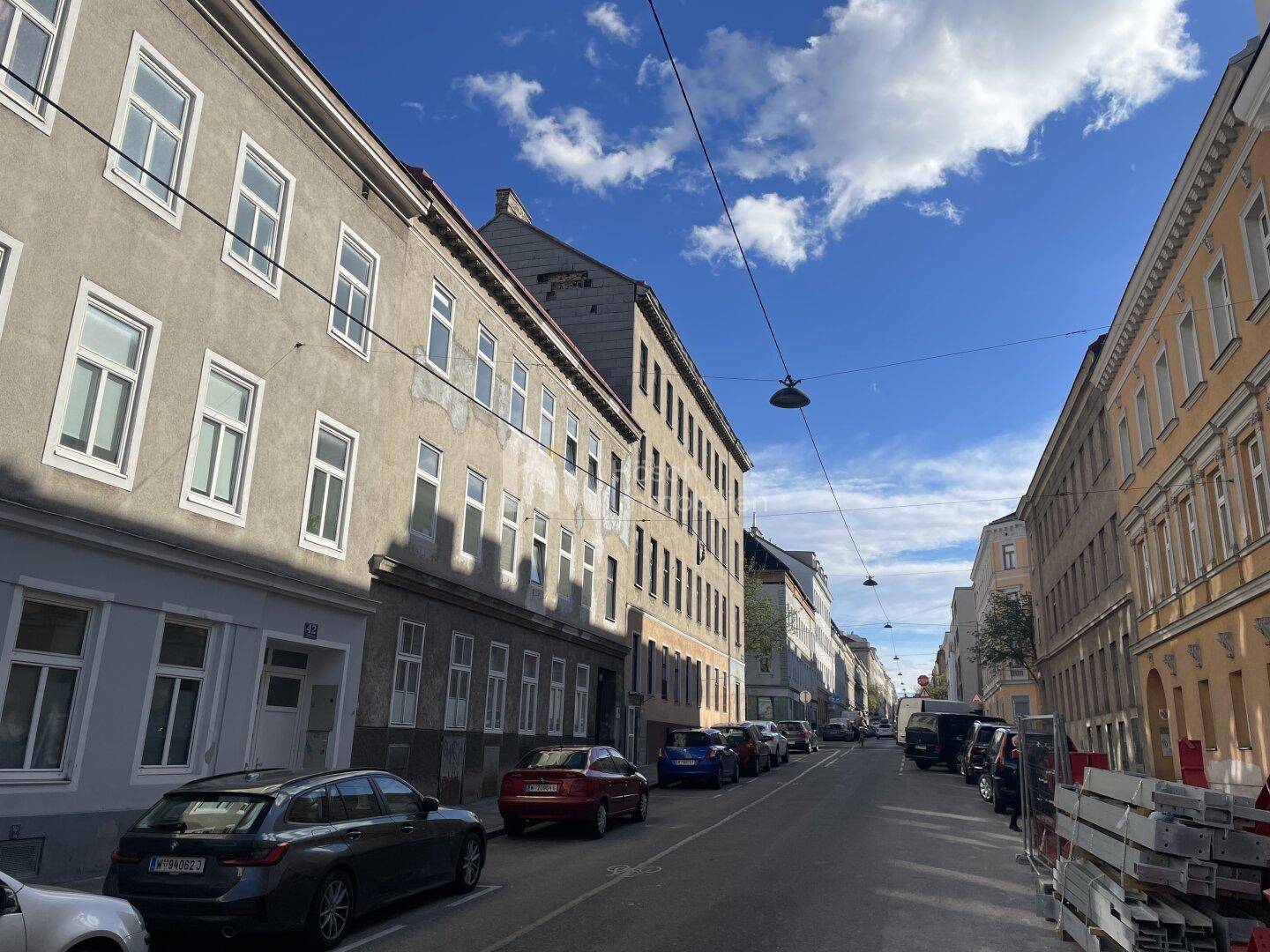 Lange Straße mit historischen Gebäuden und parkenden Fahrzeugen an einem sonnigen Tag.