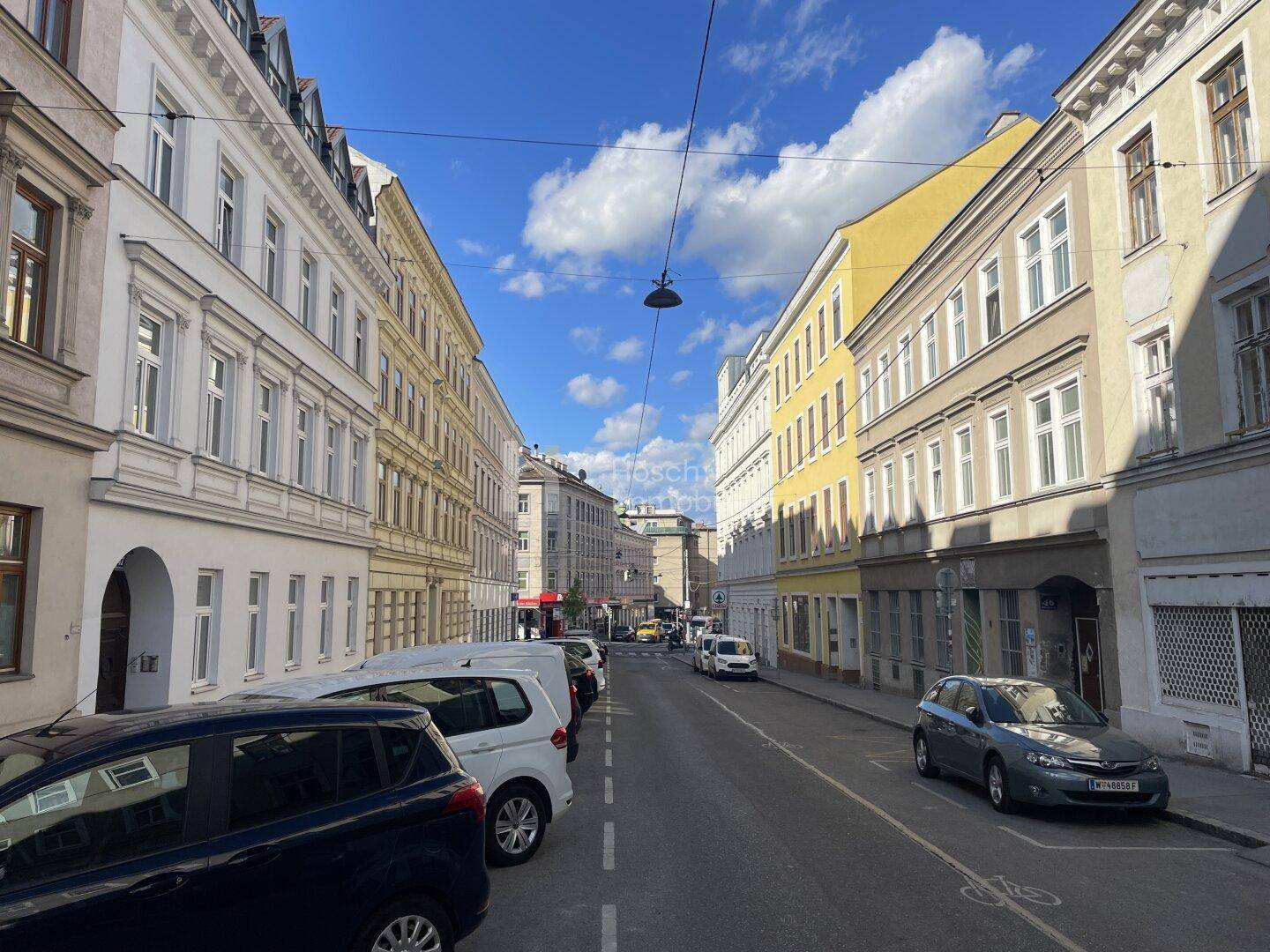 Breite Stadtstraße mit farbigen Altbaufassaden und parkenden Autos unter blauem Himmel.