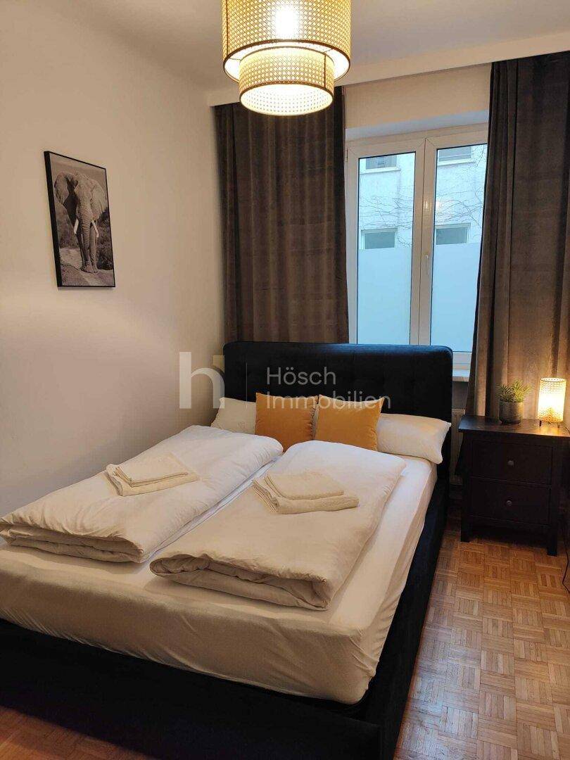 Schlafzimmer mit dunklem Bettrahmen, Nachttisch und Fenster, das Tageslicht hereinlässt.