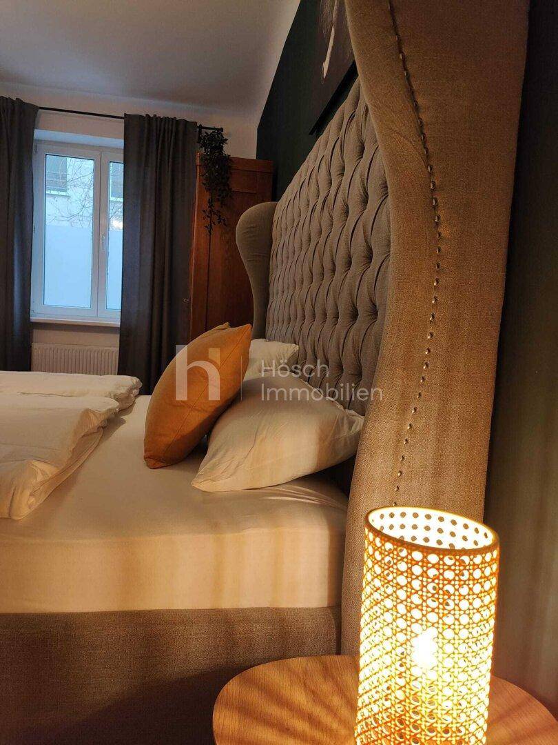 Seitenansicht des Bettes mit Nachttischlampe und Blick auf das Fenster mit Vorhängen.