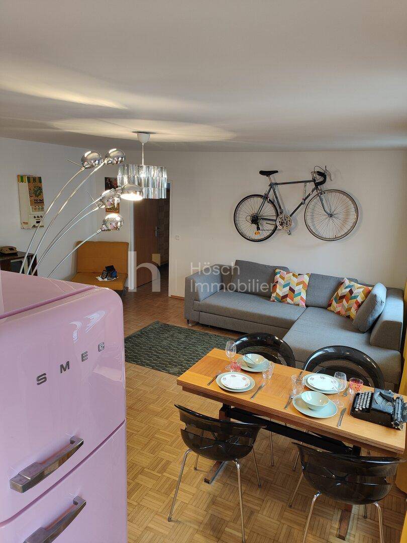Offener Wohn- und Essbereich mit Sofa, Esstisch und einem auffälligen rosa Kühlschrank.
