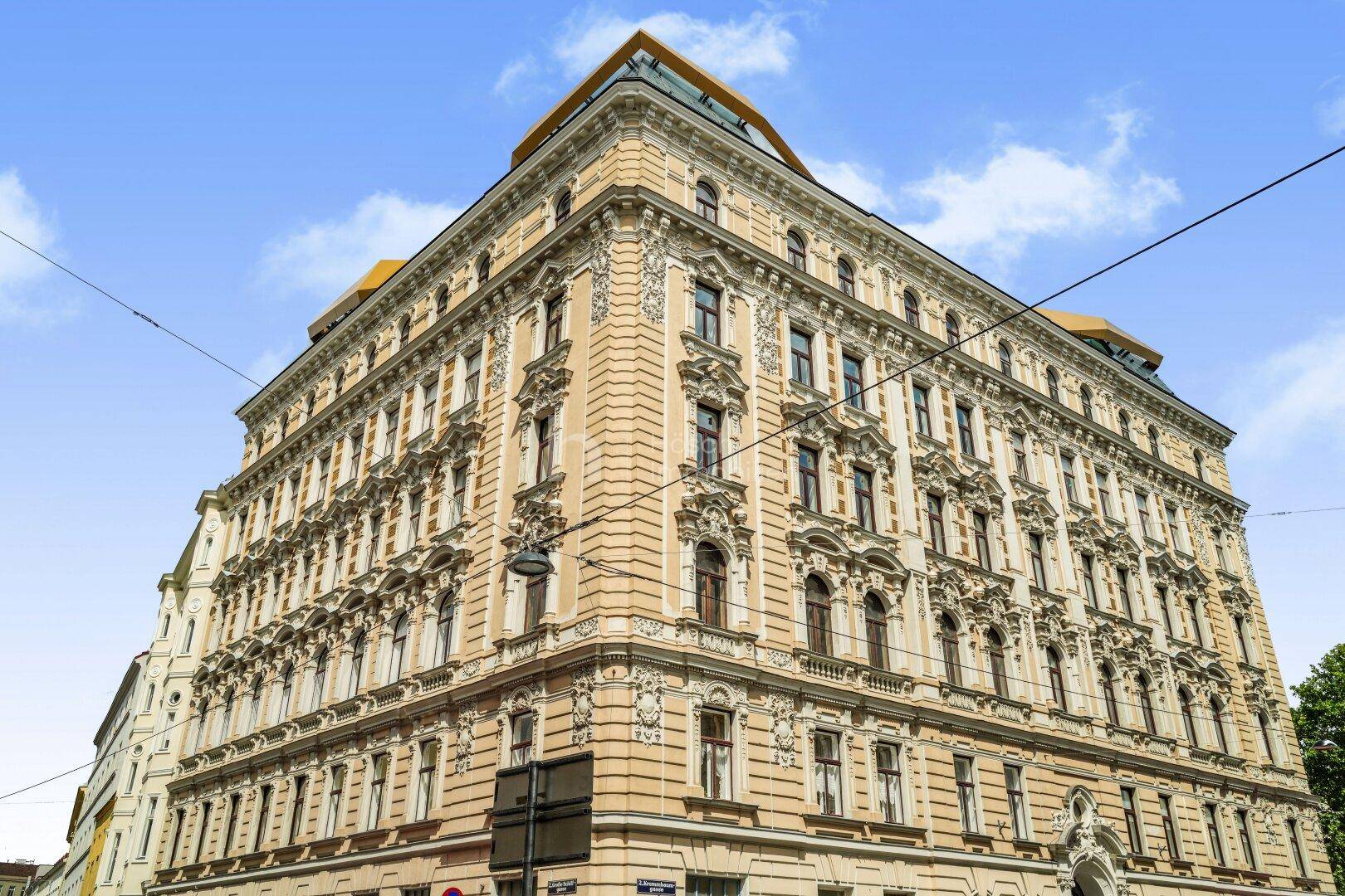 Großes Eckgebäude mit historischer, reich verzierter Fassade und moderner Dachaufstockung.