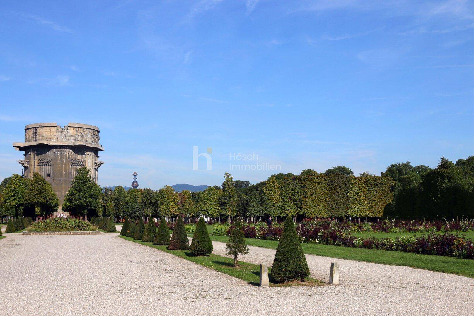 Weitläufiger Park mit Kieswegen, gepflegten Grünflächen und einem Flakturm in der Ferne.