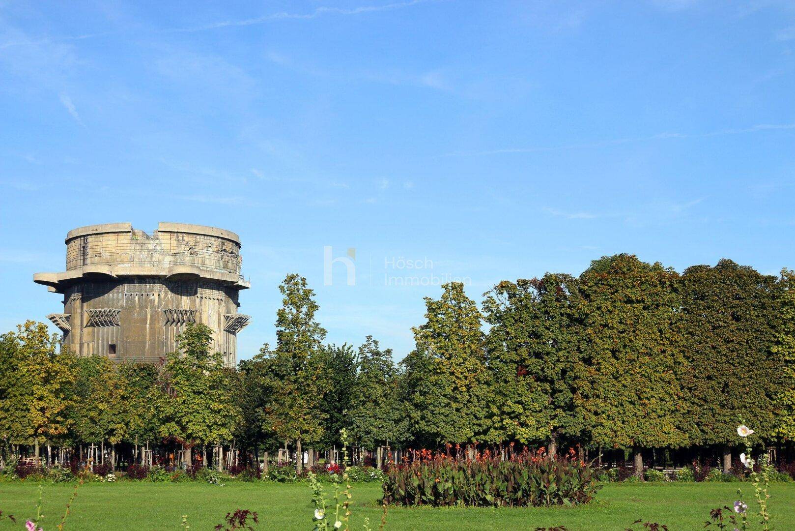 Grüne Parklandschaft mit einem historischen Flakturm im Hintergrund und hohen Bäumen.