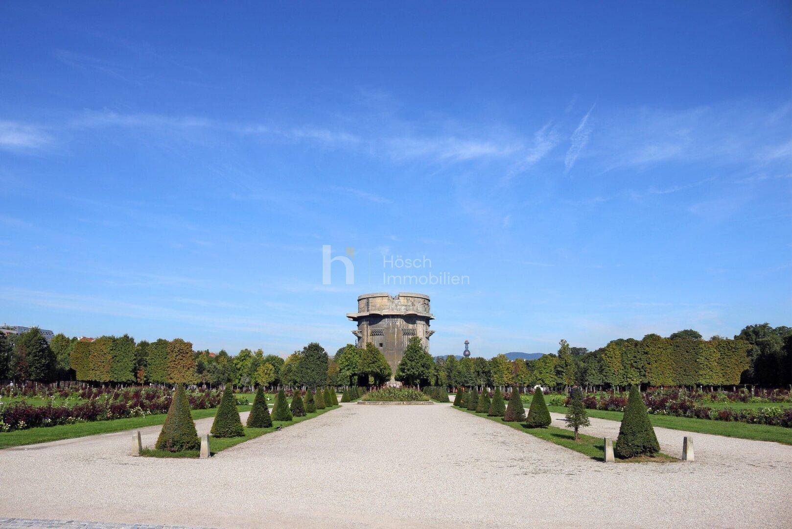 Panoramablick über einen weitläufigen Park mit symmetrischen Wegen und einem Flakturm.