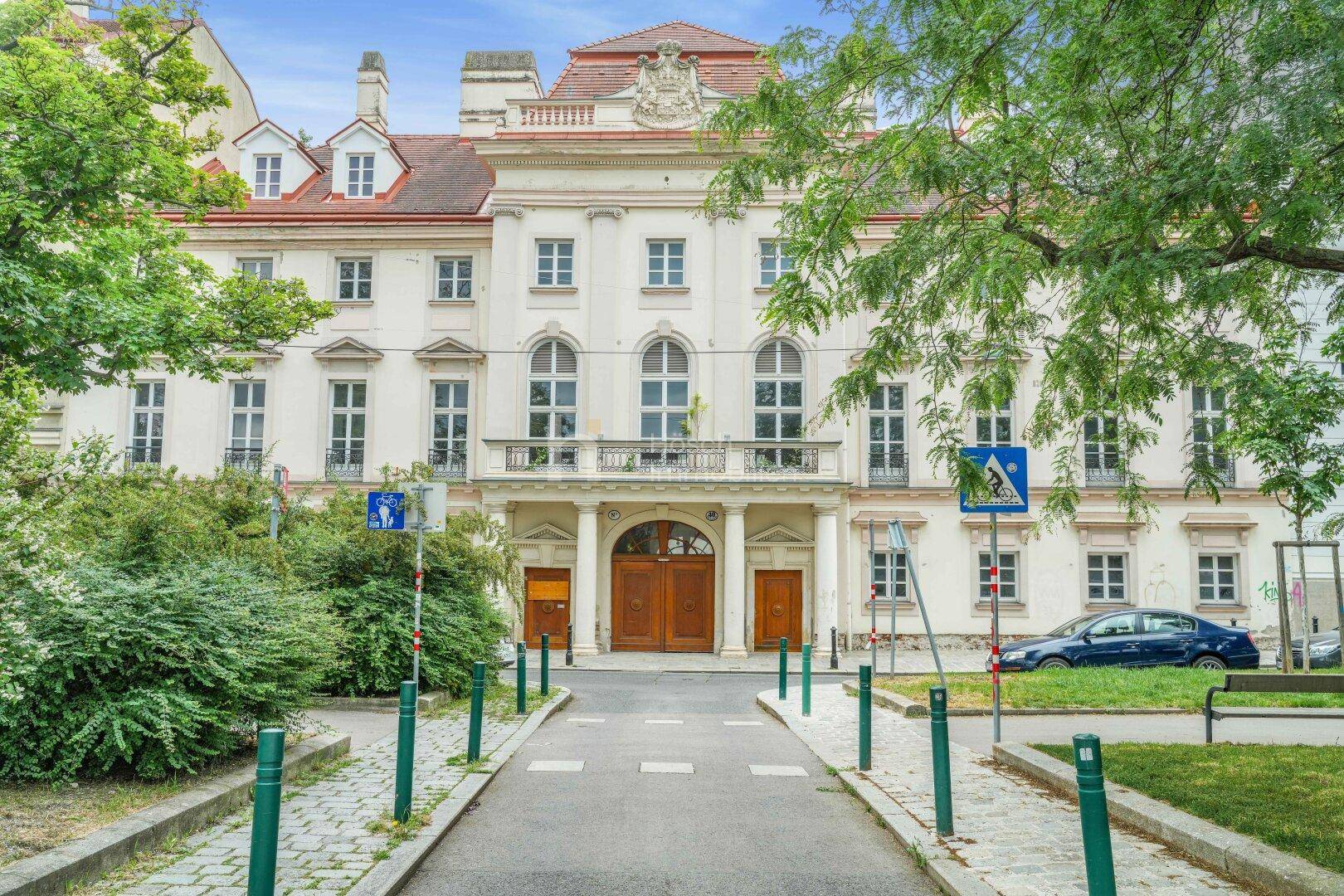 Historisches Gebäude mit klassischer Architektur, umgeben von Grünflächen und Bäumen in urbaner Umgebung.