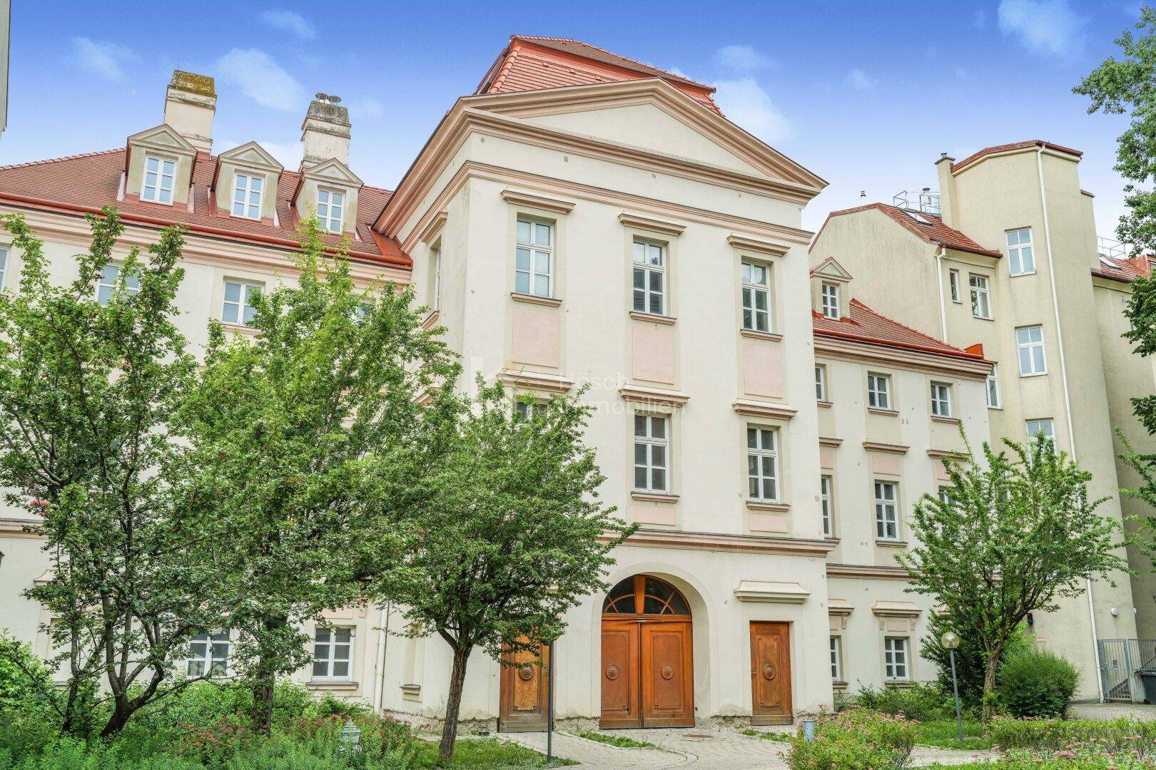 Historisches Gebäude mit klassischer Architektur, umgeben von kleinen Grünflächen und Bäumen.