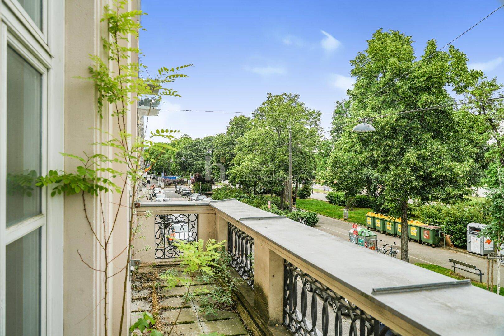 Balkon mit schmiedeeisernem Geländer und Blick auf eine von Bäumen gesäumte Straße.