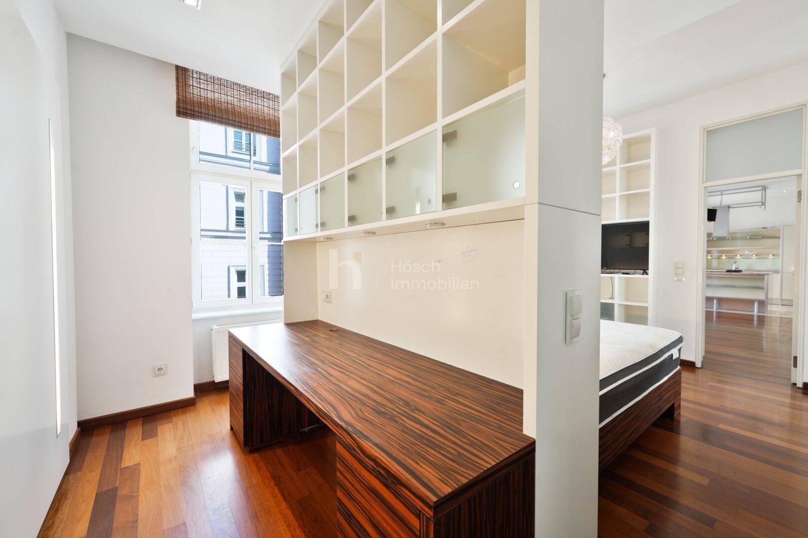 Arbeitsbereich mit großem Holzschreibtisch und integriertem Regal, angrenzend an ein Schlafzimmer.