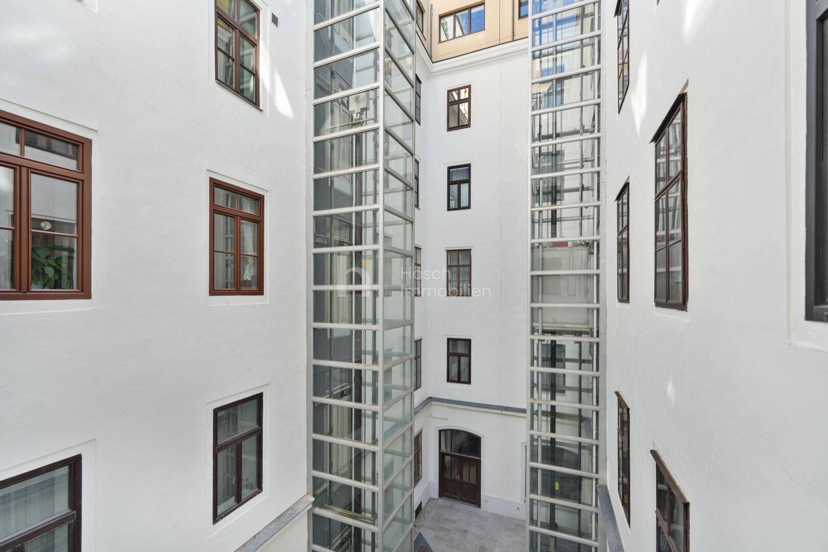 Innenhof eines Gebäudes mit zwei modernen Glasaufzügen und weiß gestrichenen Wänden.