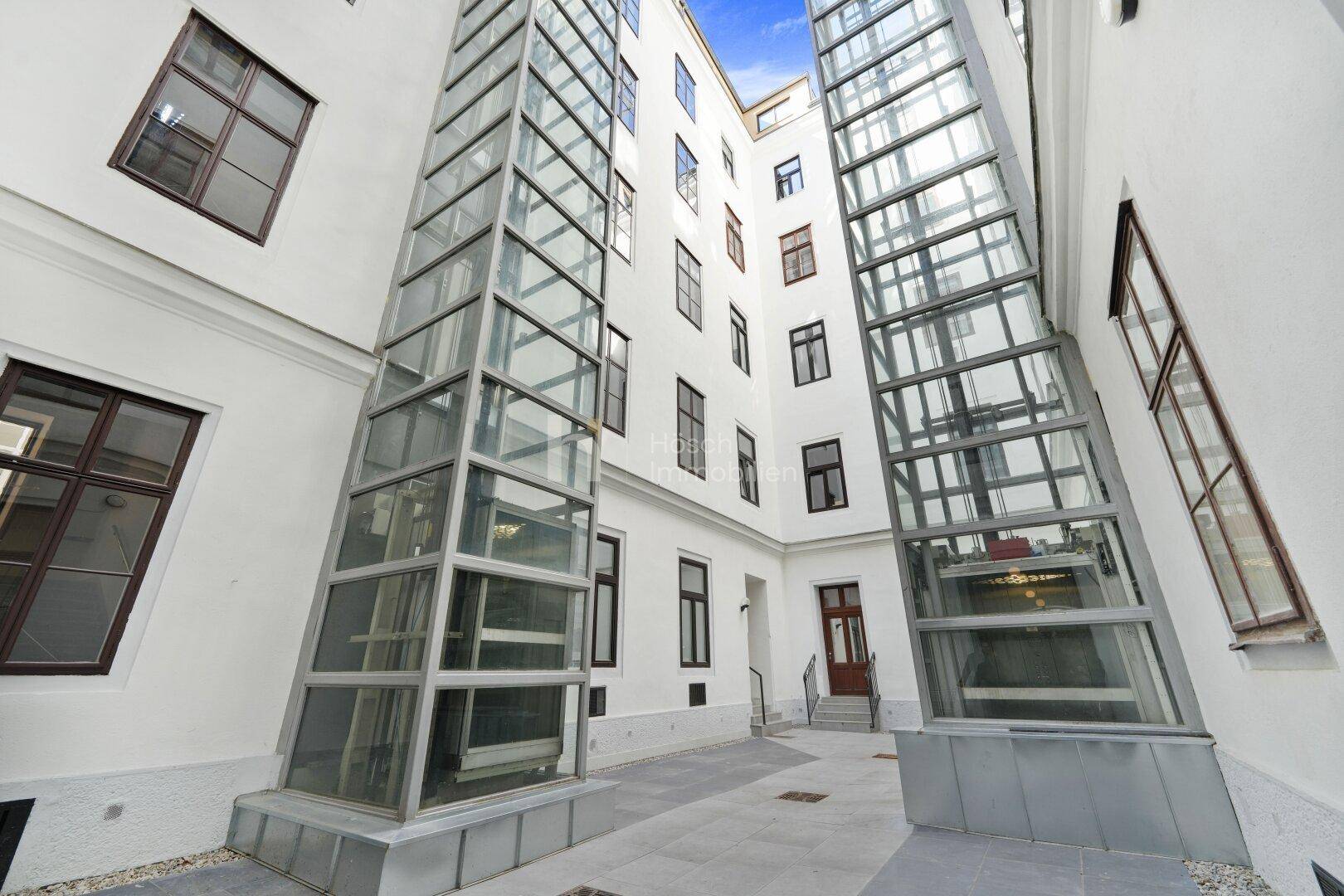 Heller Innenhof mit zwei Glasaufzügen und klassischer Architektur im Hintergrund.