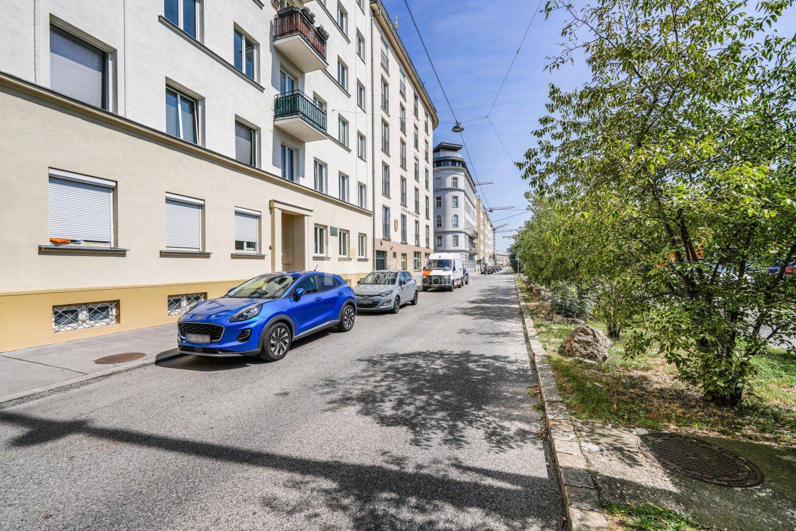 Ruhige Wohnstraße mit geparkten Autos und der Fassade des Mehrfamilienhauses.