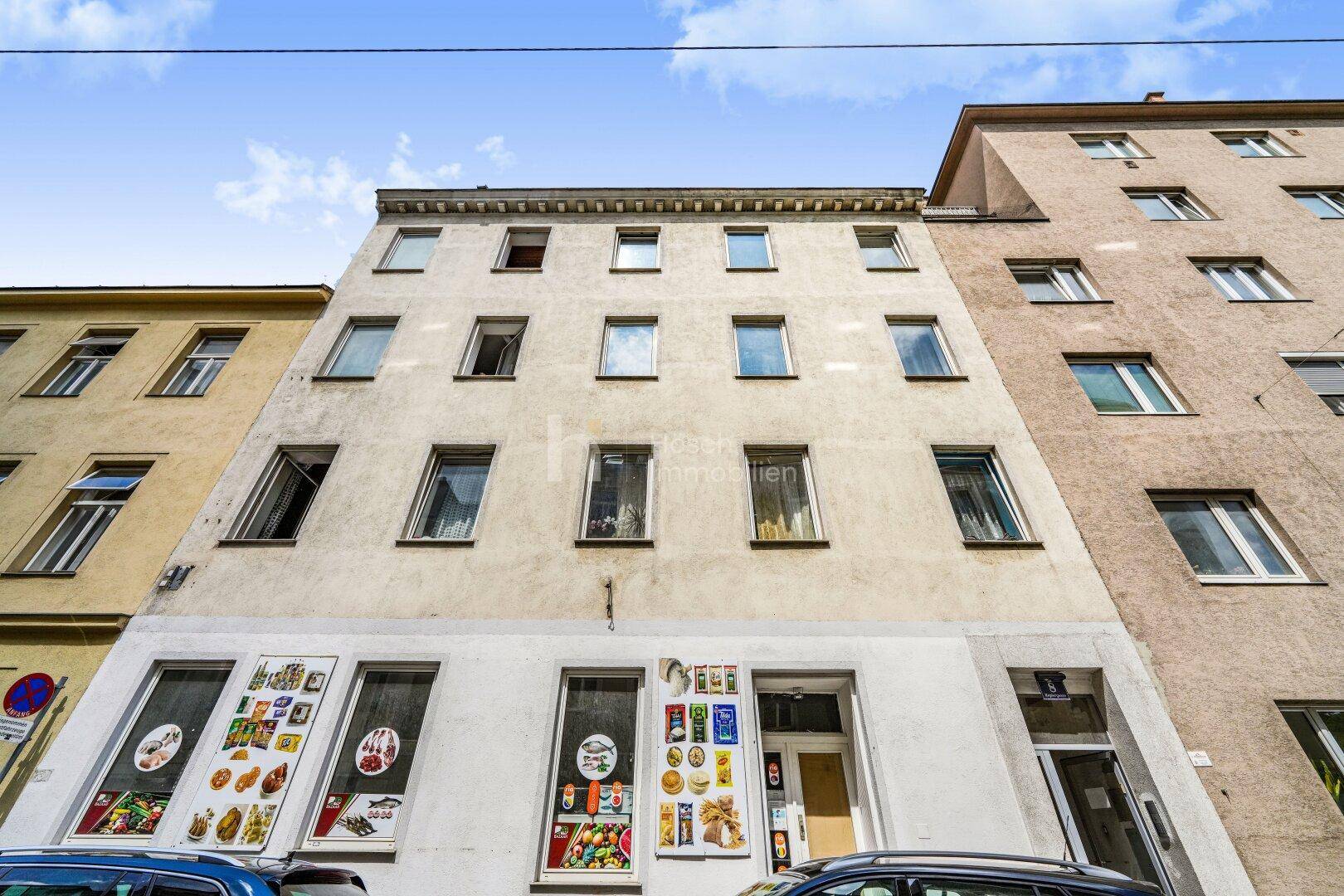 Fassade eines Mehrfamilienhauses mit Geschäften im Erdgeschoss unter blauem Himmel.