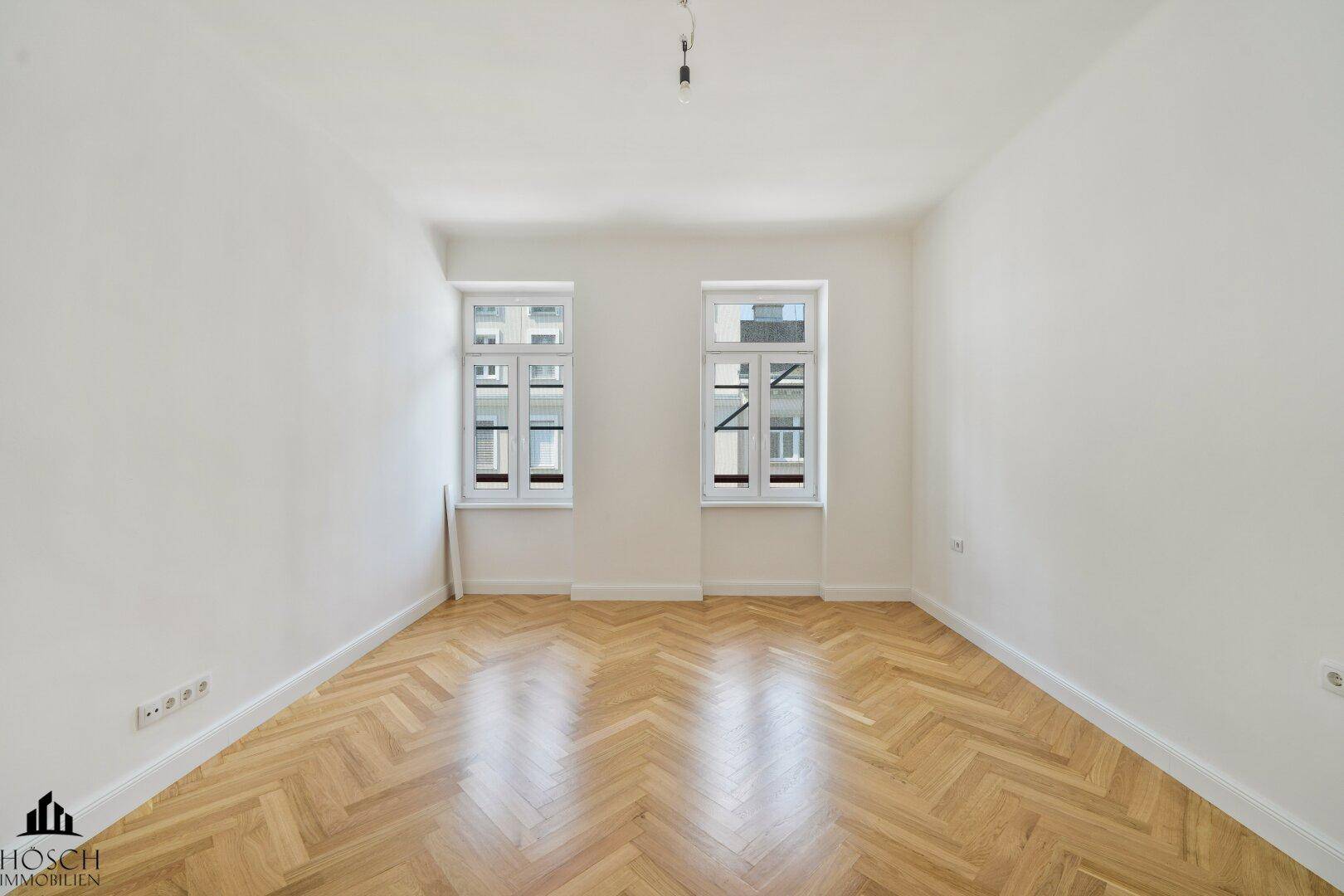 Leerer Raum mit Fischgrätparkett und zwei hohen Fenstern, ideal für individuelle Gestaltung.