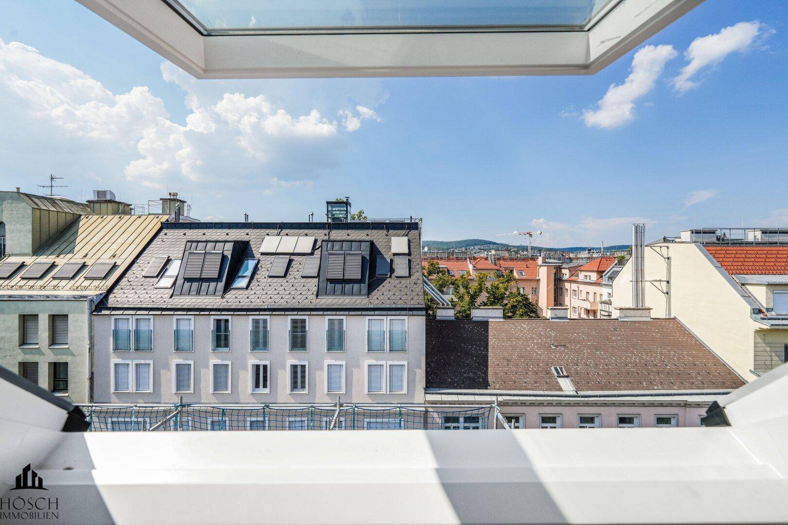 Weitblick über die Dächer der Stadt mit blauem Himmel durch ein Dachfenster.