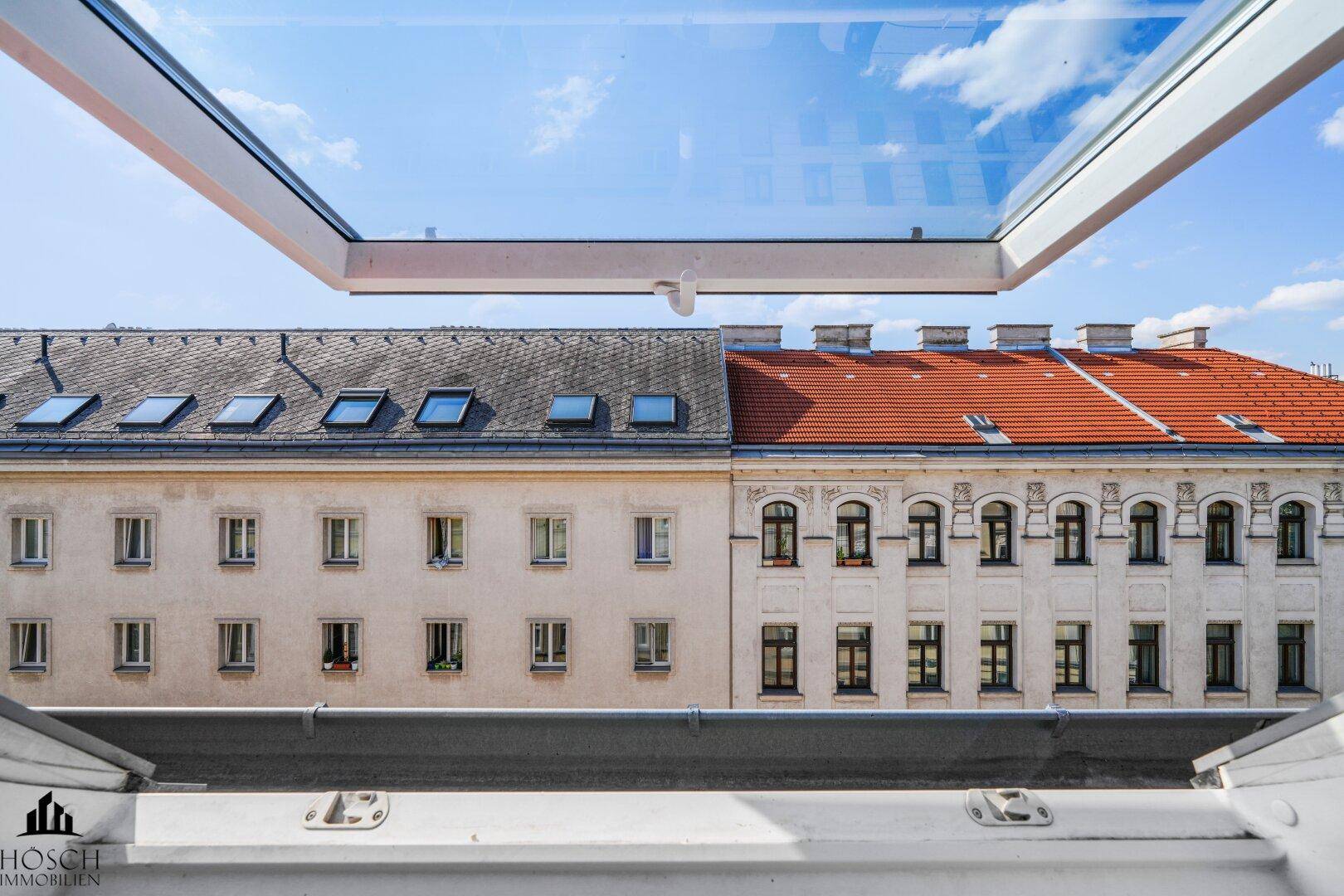 Blick aus einem Dachfenster auf gegenüberliegende Gebäude und den blauen Himmel.
