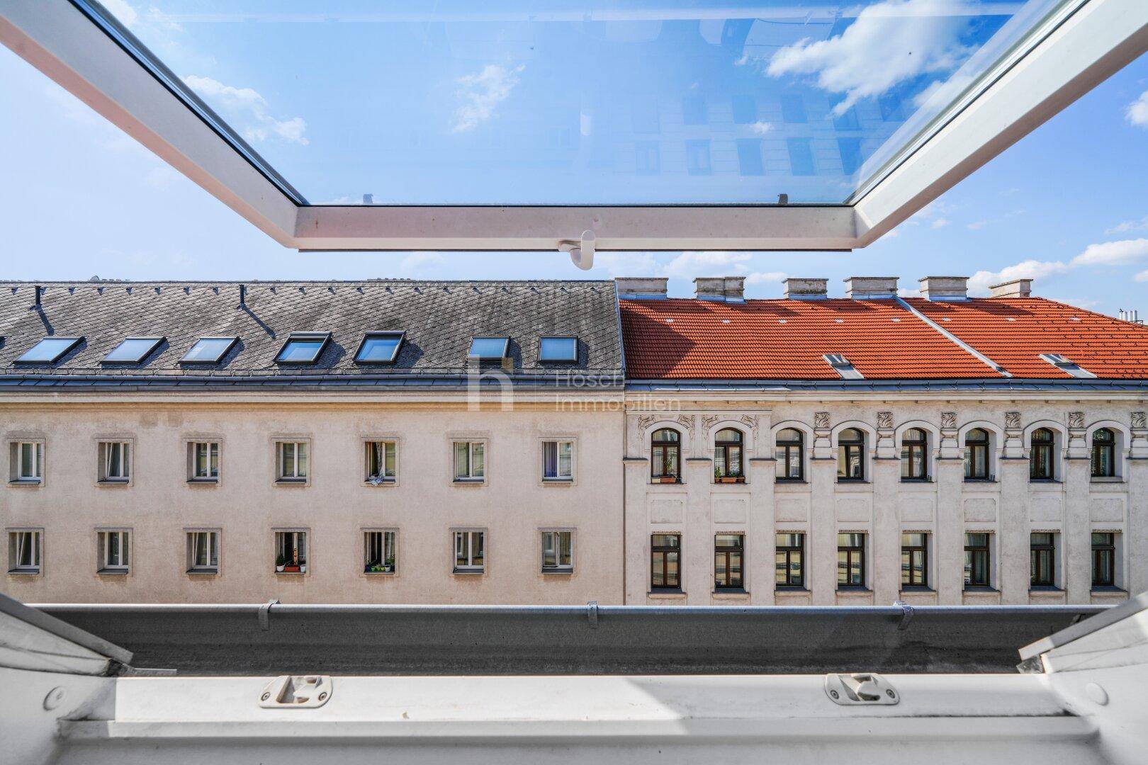 Blick aus einem geöffneten Dachfenster auf die Dächer benachbarter Gebäude und den blauen Himmel.