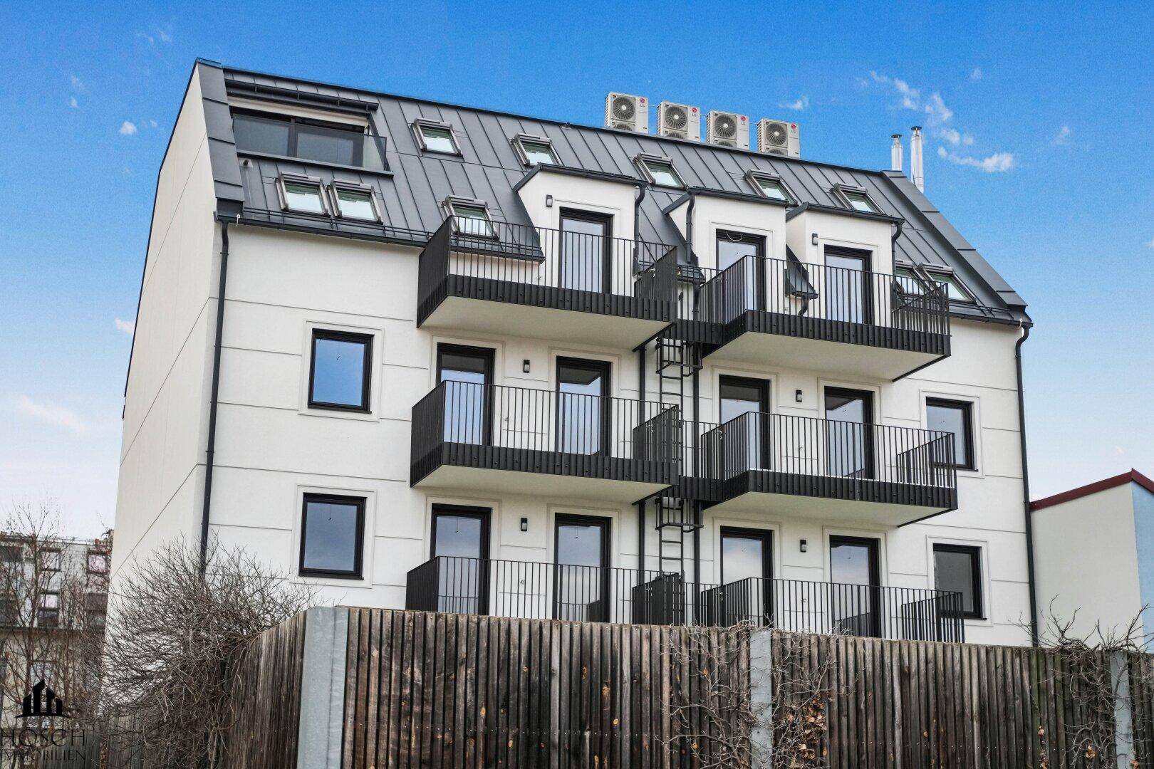 Rückseite des modernen Gebäudes mit mehreren Balkonen, Dachfenstern und Klimaanlagen auf dem Dach.