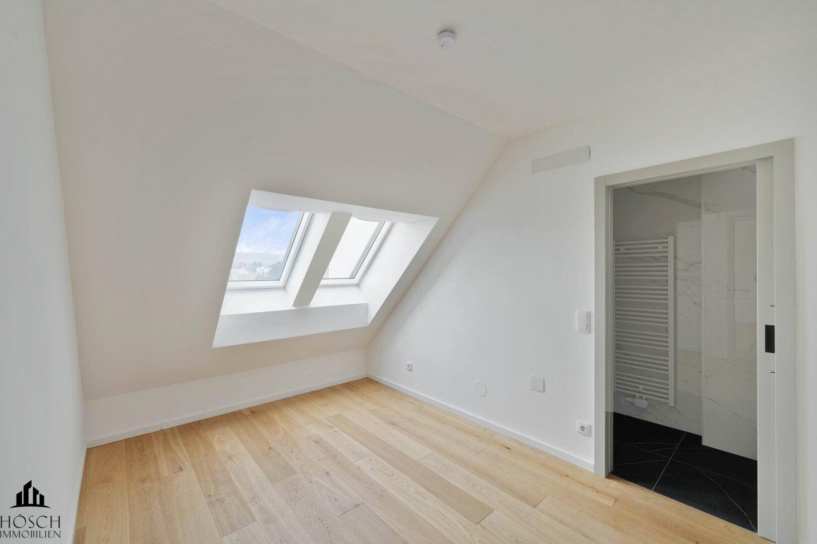 Heller, leerer Raum mit Holzboden, Dachschrägen, Dachfenstern und Zugang zu einem Badezimmer.