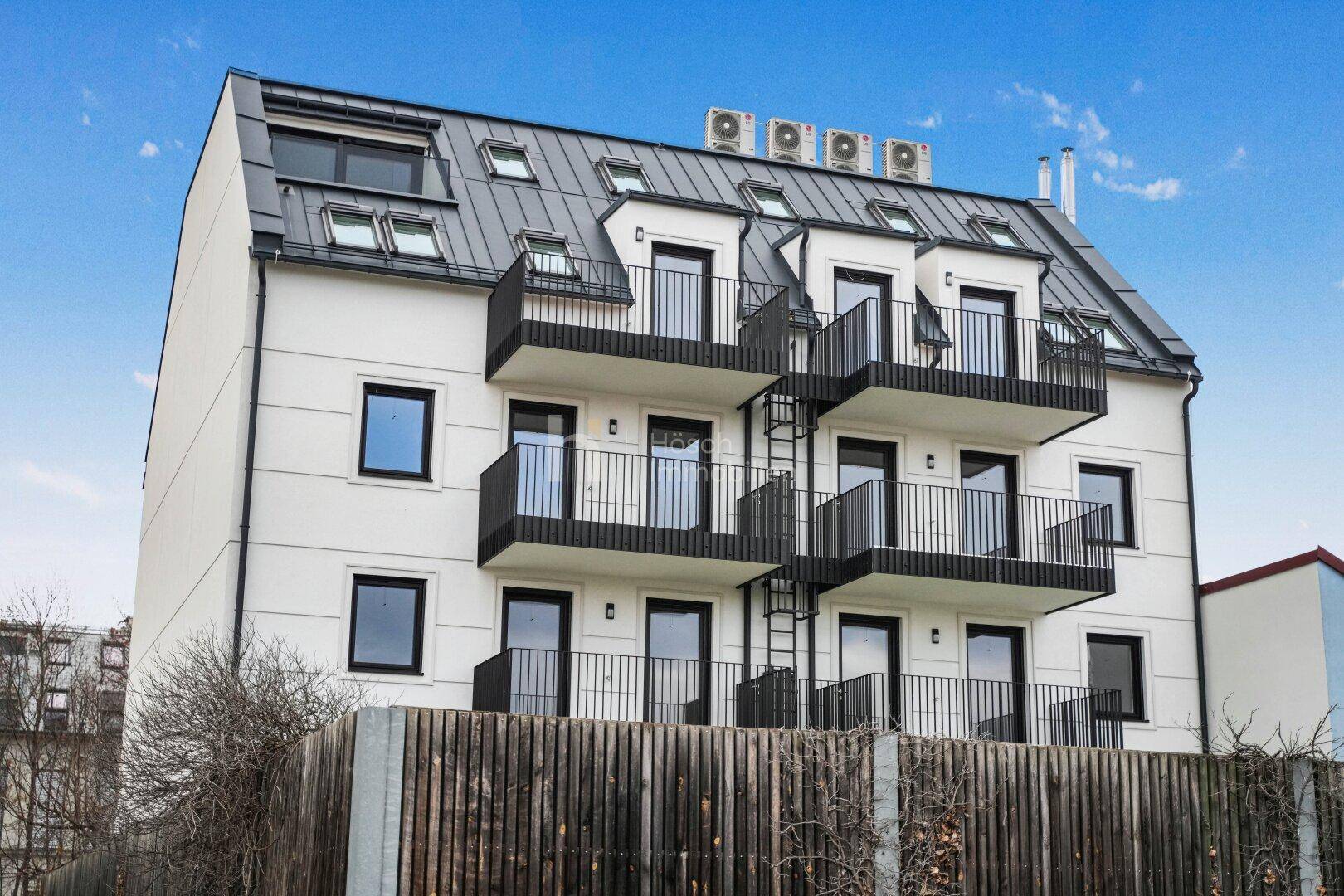 Moderne Wohnfassade mit mehreren Balkonen und Dachfenstern unter einem Mansarddach.