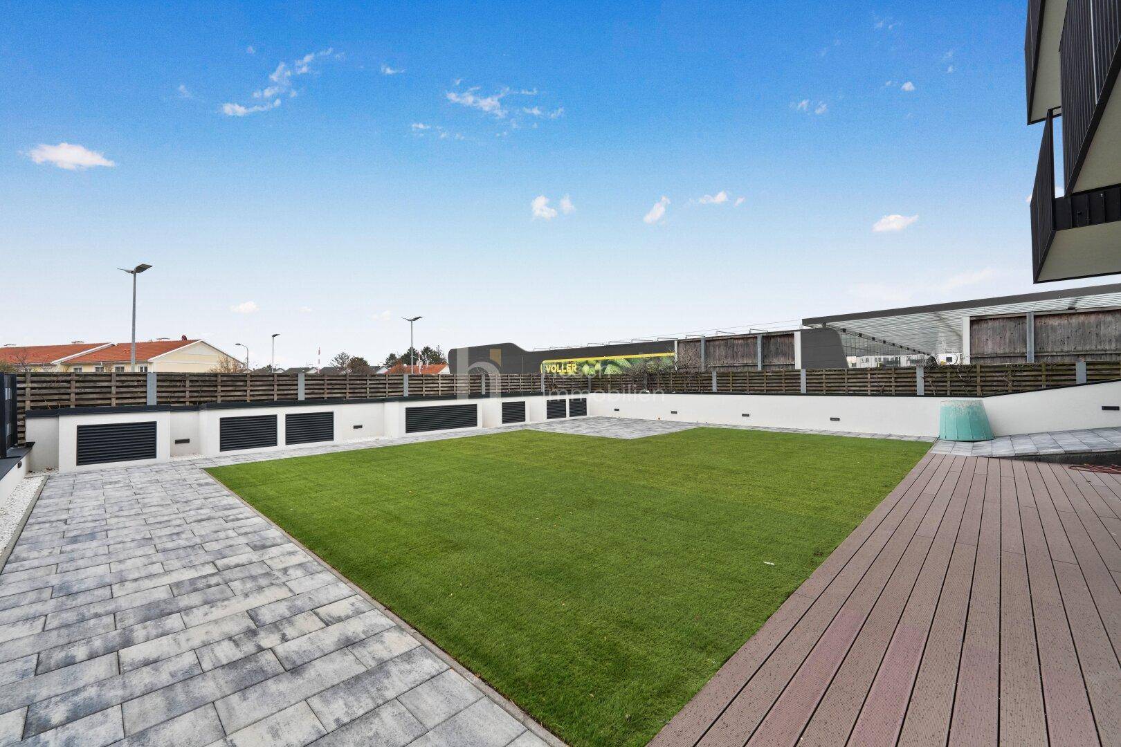 Großer, gepflegter Gartenbereich mit Rasen und Holzterrasse, umgeben von einer modernen Wohnanlage.
