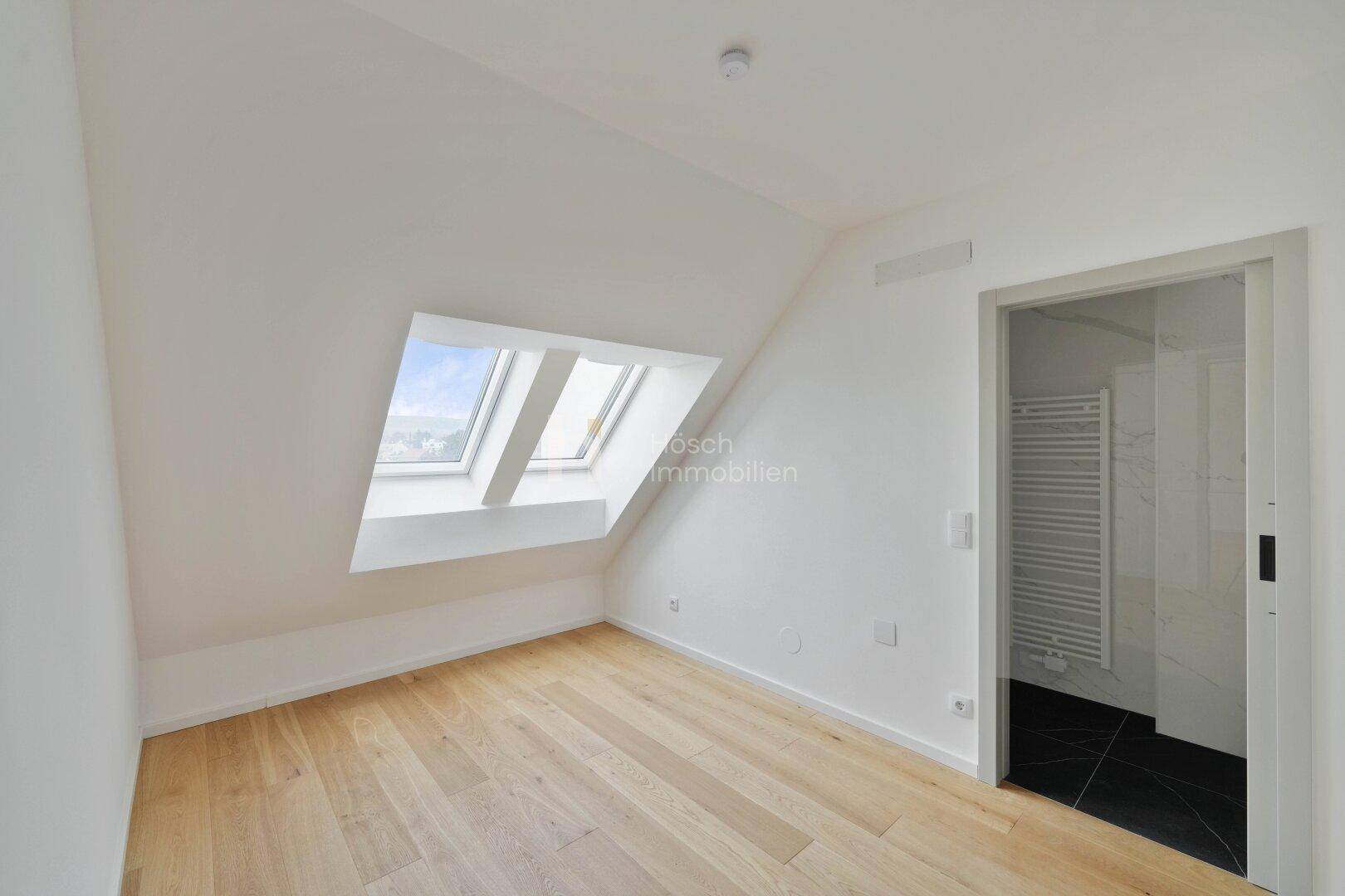 Helles Schlafzimmer im Dachgeschoss mit Holzboden, Dachfenstern und Zugang zu einem Badezimmer.