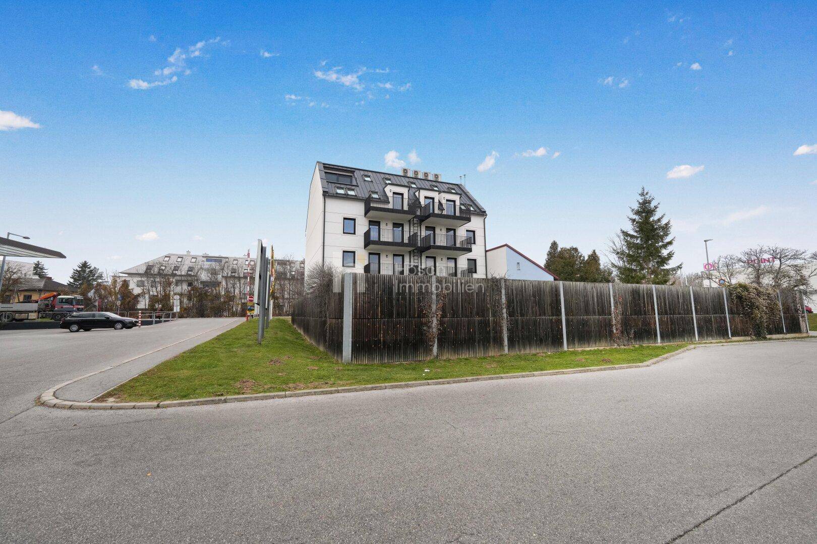 Modernes Mehrfamilienhaus mit Balkonen und dunklem Dach, umgeben von einer Straße und Grünflächen.