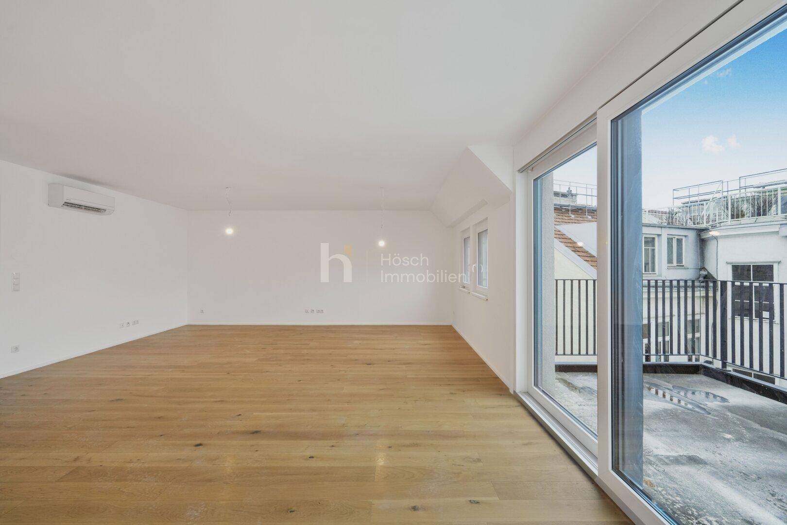 Geräumiger, leerer Wohnbereich mit Holzboden, großen Fenstern und Zugang zu einer Terrasse.