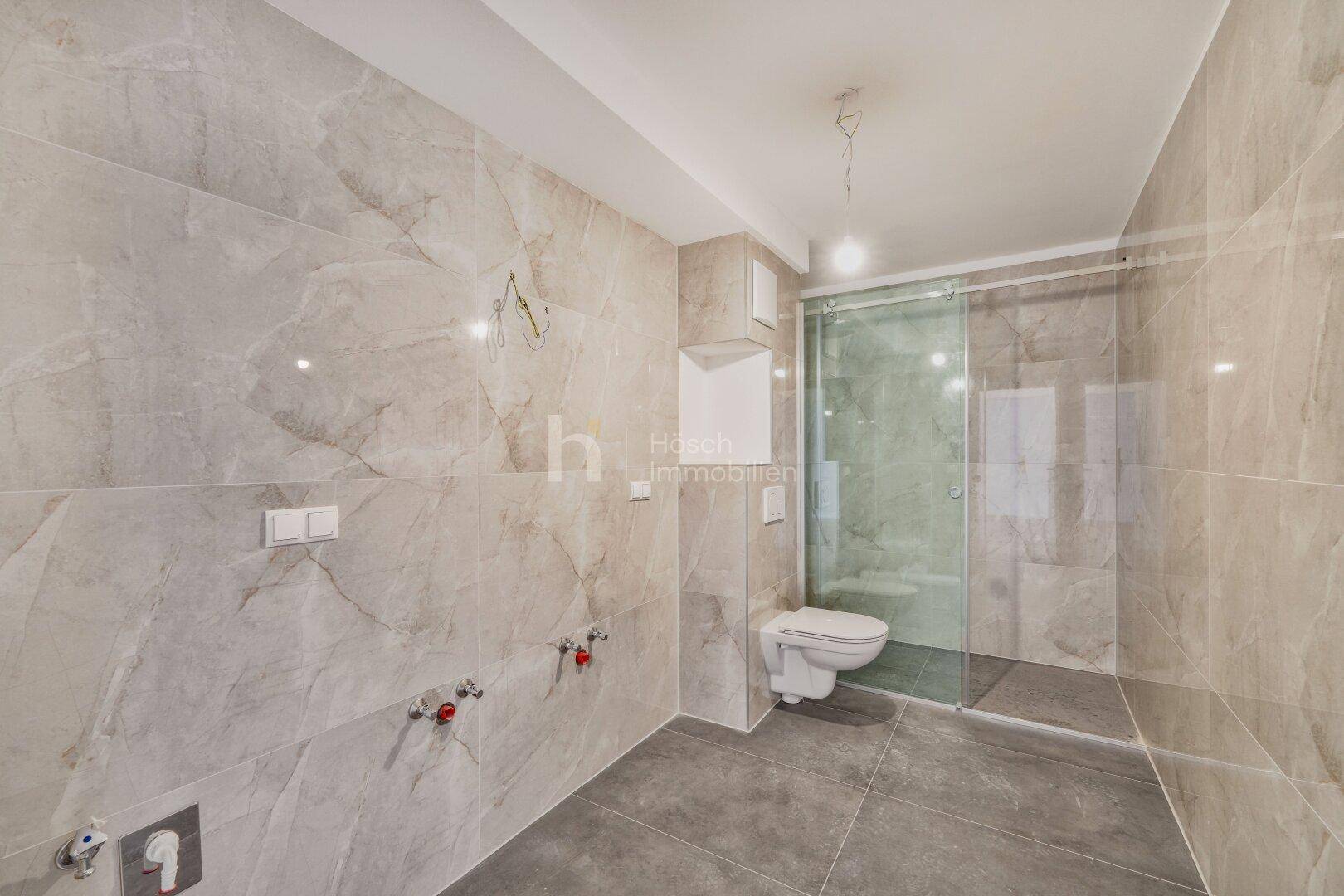 Modernes Badezimmer mit großformatigen grauen Fliesen, wandhängendem WC und ebenerdiger Dusche.