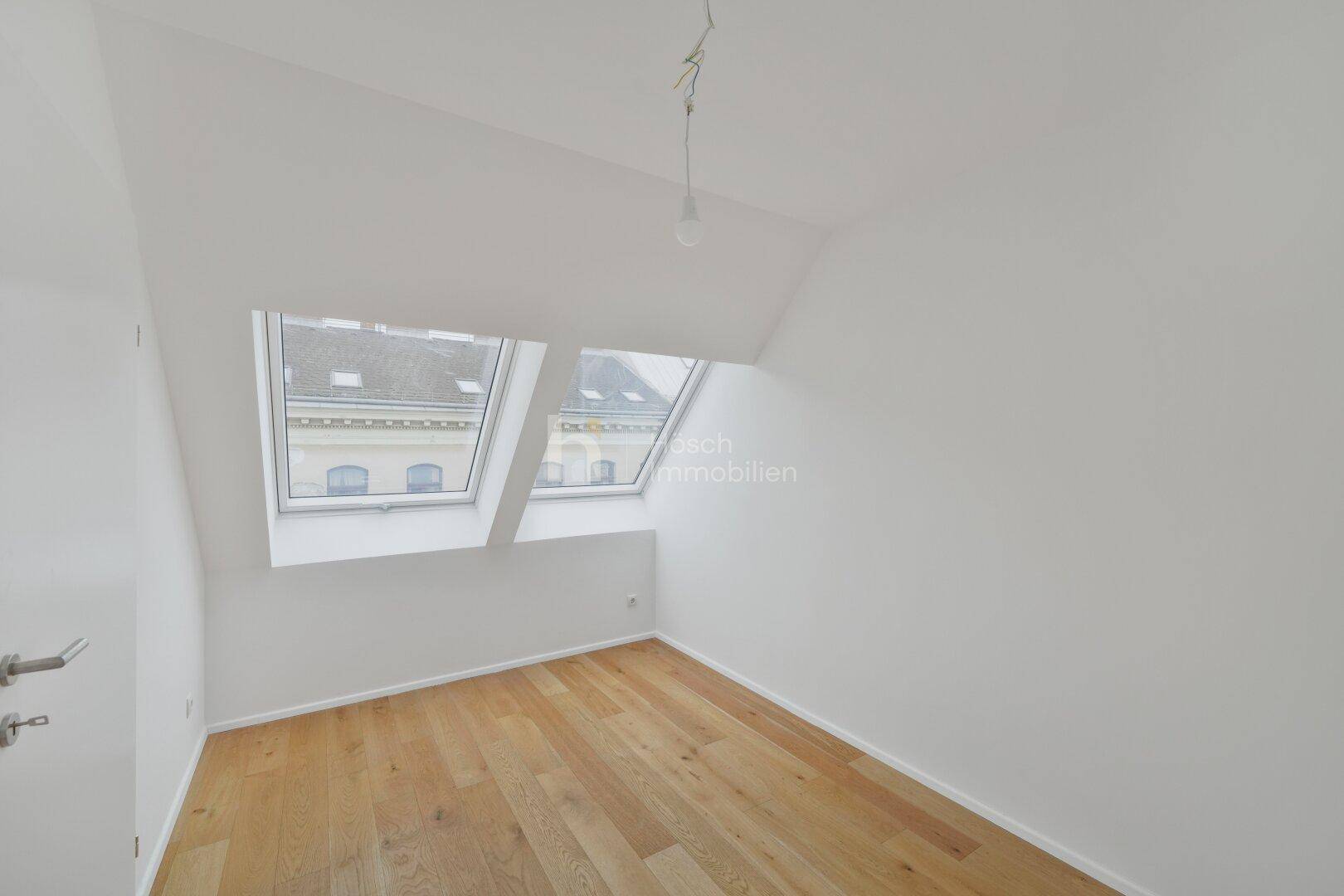 Helles Zimmer im Dachgeschoss mit schrägen Wänden, Holzfußboden und zwei großen Dachfenstern.