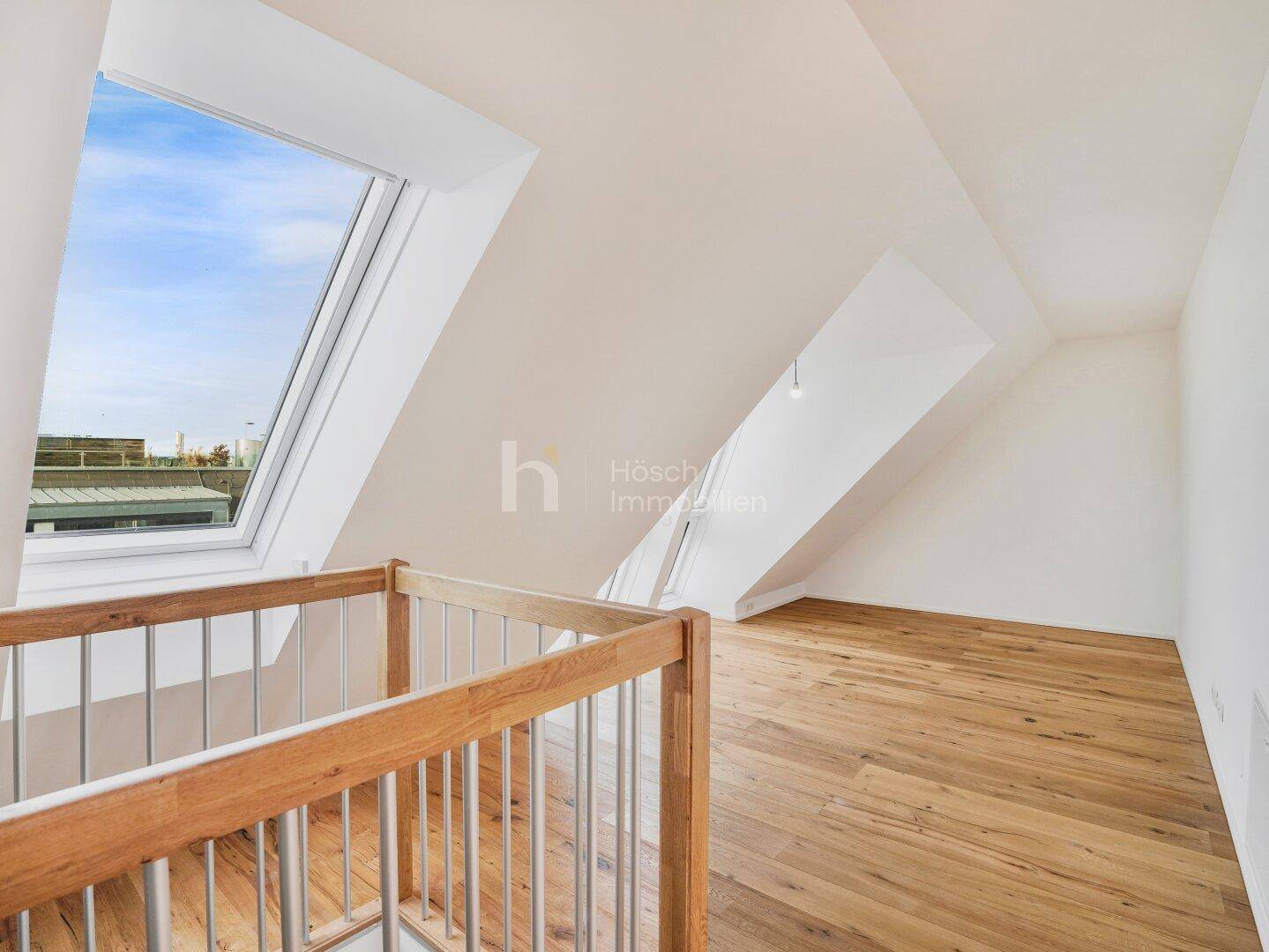 Geräumiger Dachgeschossraum mit Holzboden, Dachfenster und Blick auf die Umgebung, inklusive Treppengeländer.