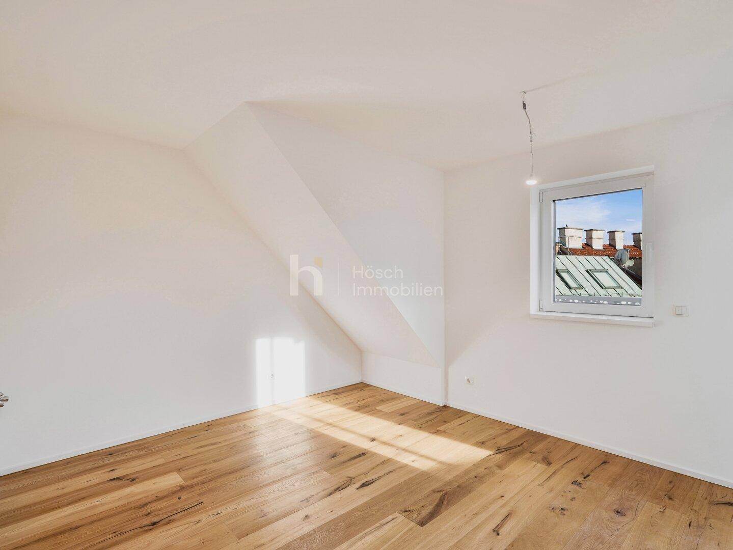 Helles Zimmer im Dachgeschoss mit Holzboden und einem Fenster, das viel Sonnenlicht hereinlässt.