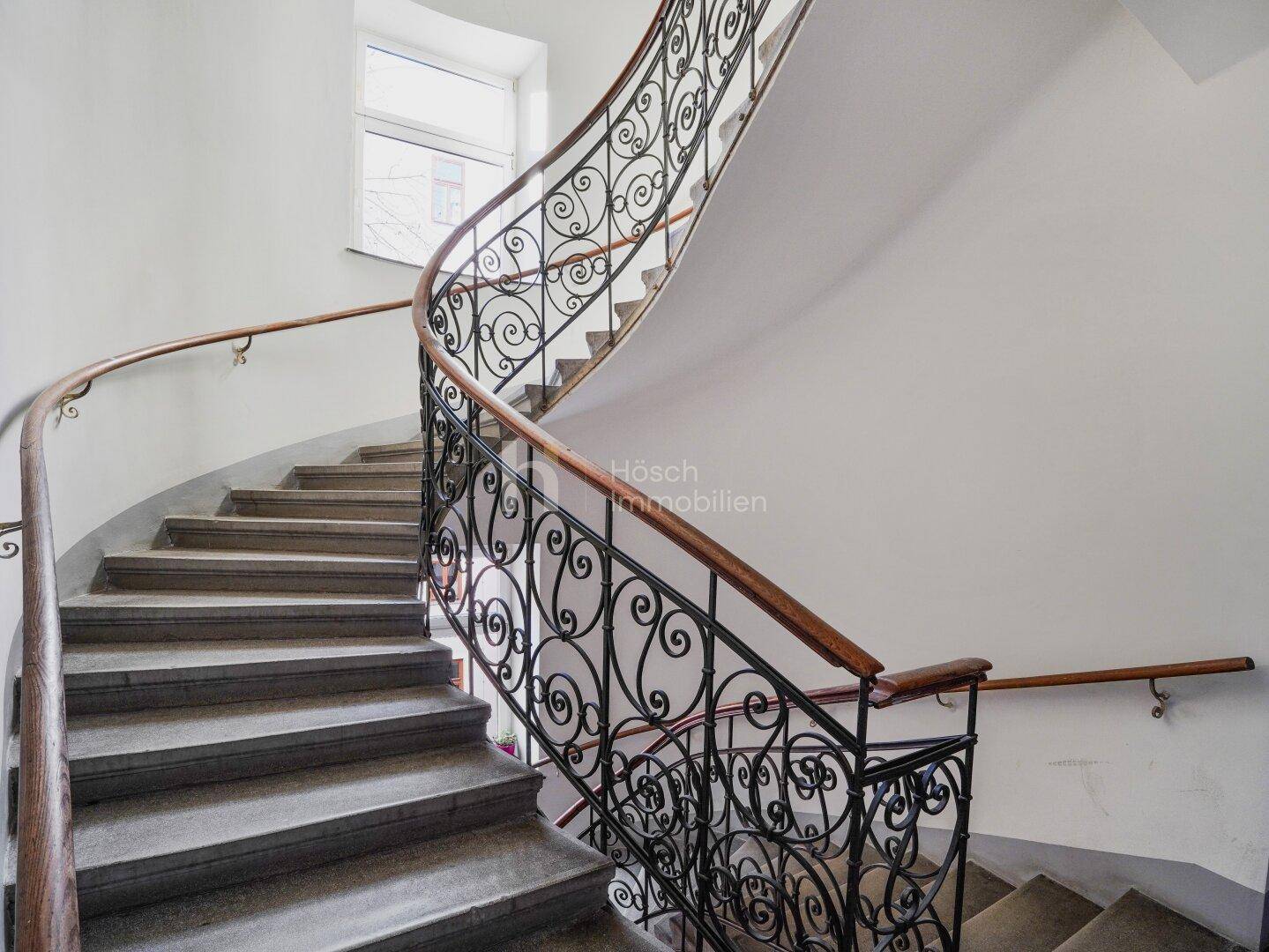 Elegantes Treppenhaus mit geschwungenem Geländer und kunstvollen Verzierungen, typisch Altbau.